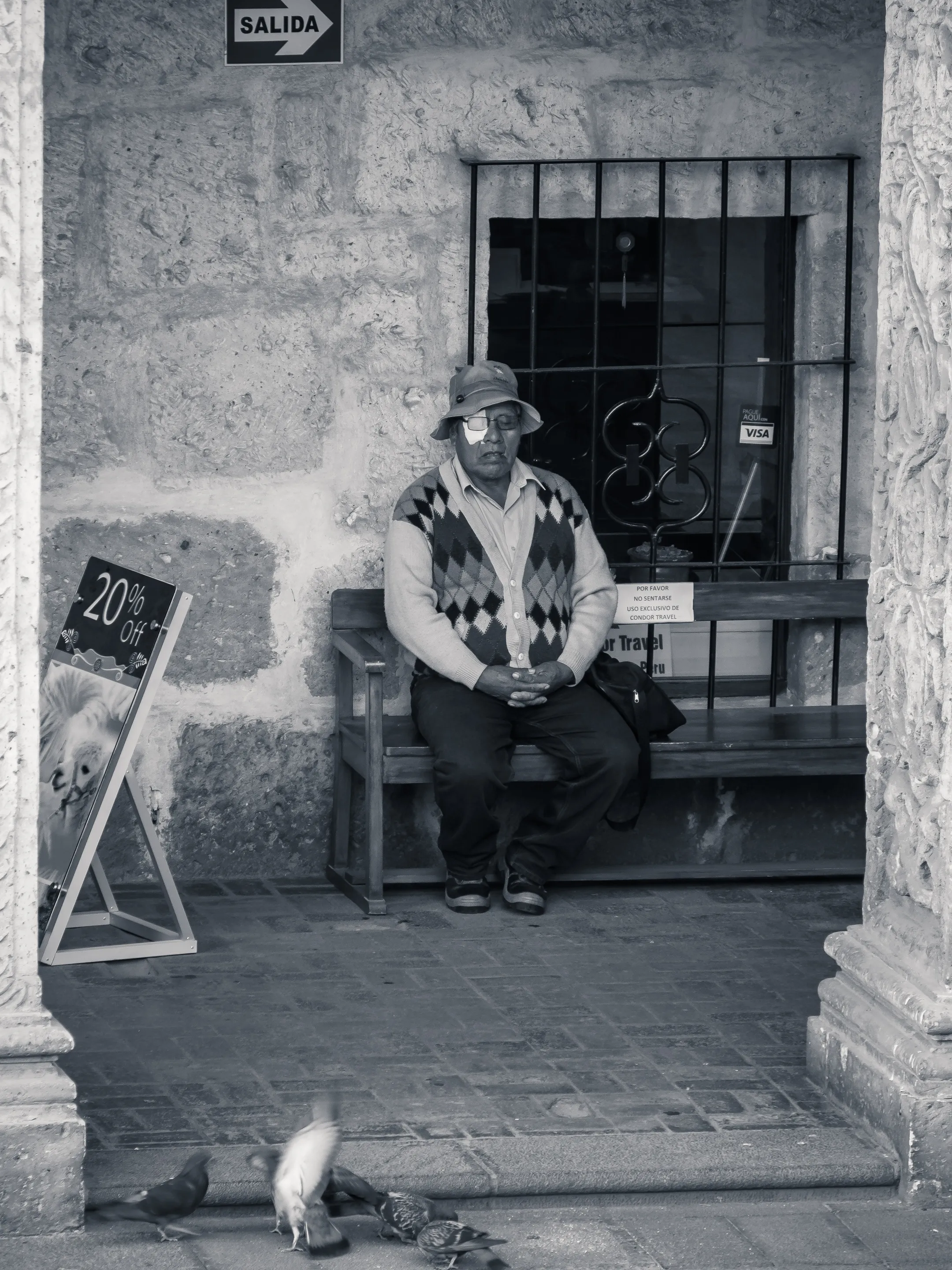 Man in hat and sweater sitting on bench outside, feeding birds, sign "SALIDA" above.