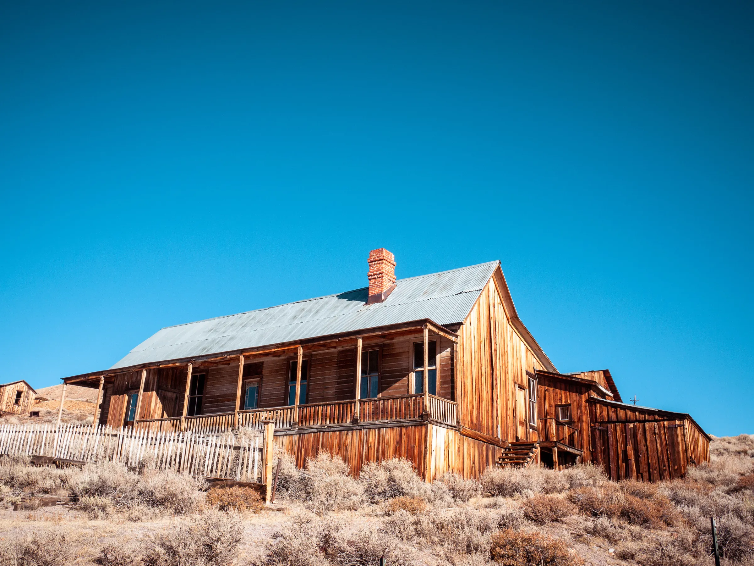 Weathered wooden house with porch, surrounded by dry grass under clear blue sky