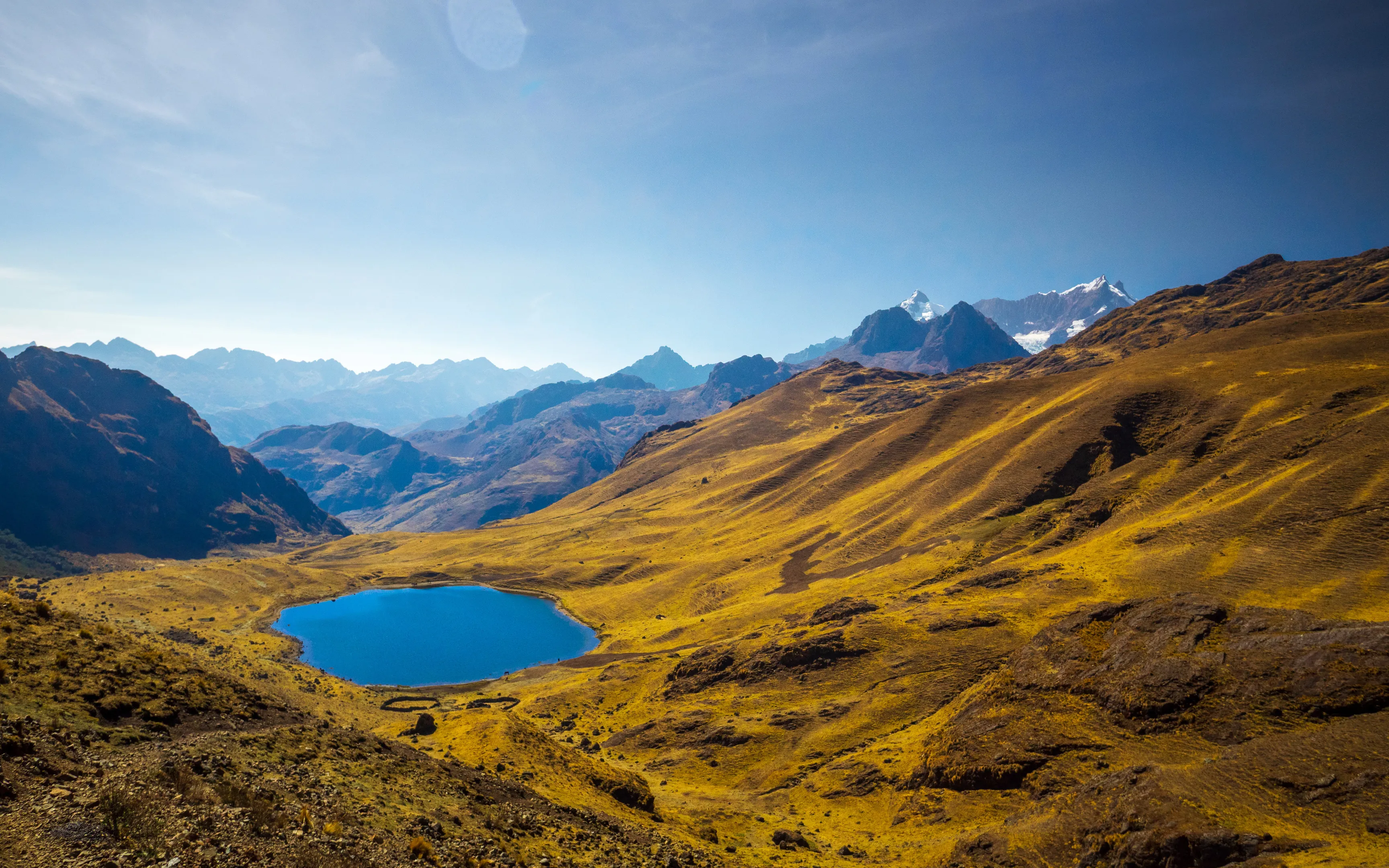 Mountainous landscape with a deep blue lake in the foreground. Golden hills roll towards snow-capped peaks under a bright sky.