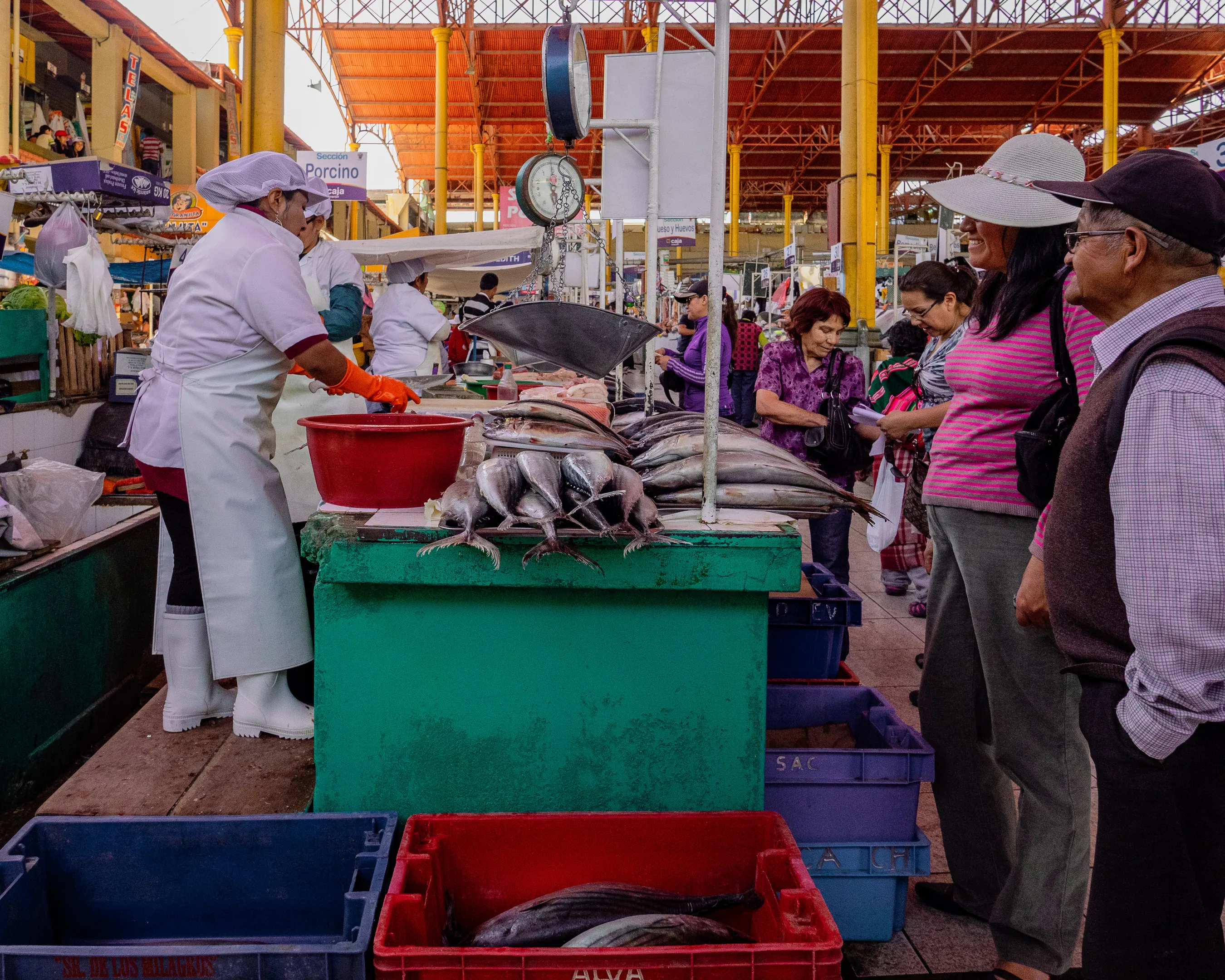 A bustling fish market with vendors in white uniforms attending to customers, various fish displayed in bins.