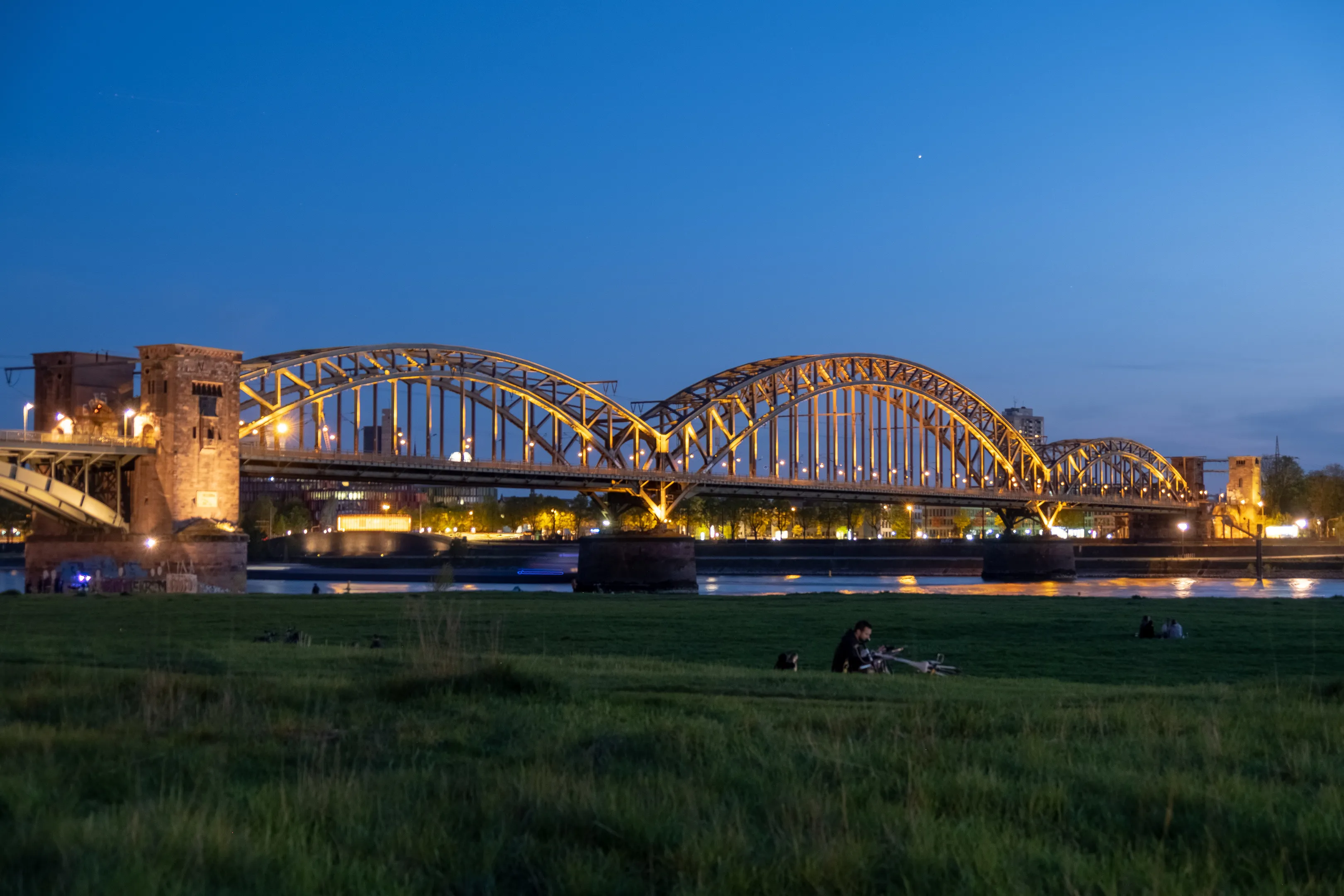 Lit bridge over river at dusk with people picnicking in foreground, grass, clear sky.