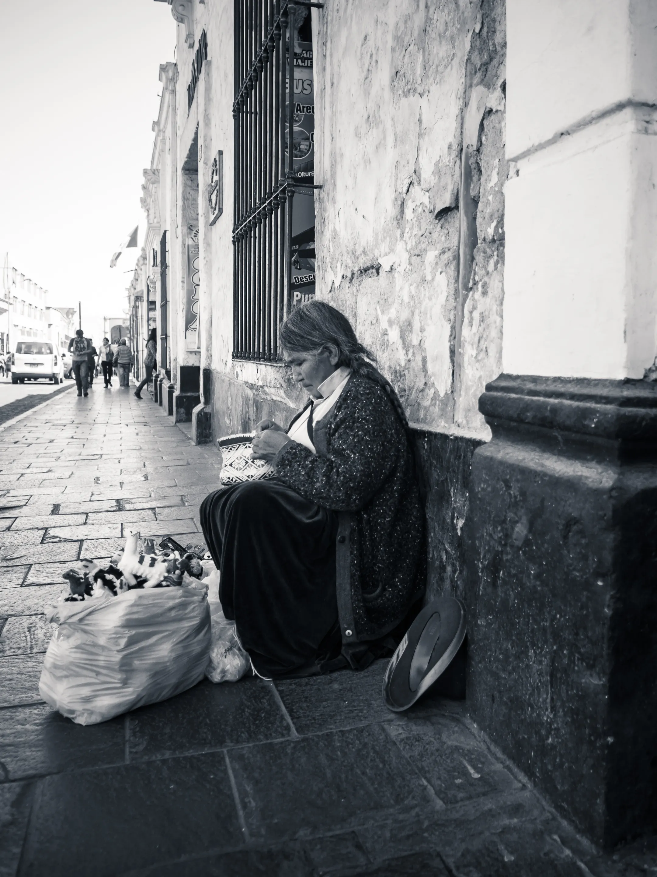 Woman sitting against wall on street, holding bread, with a bag of items beside her.