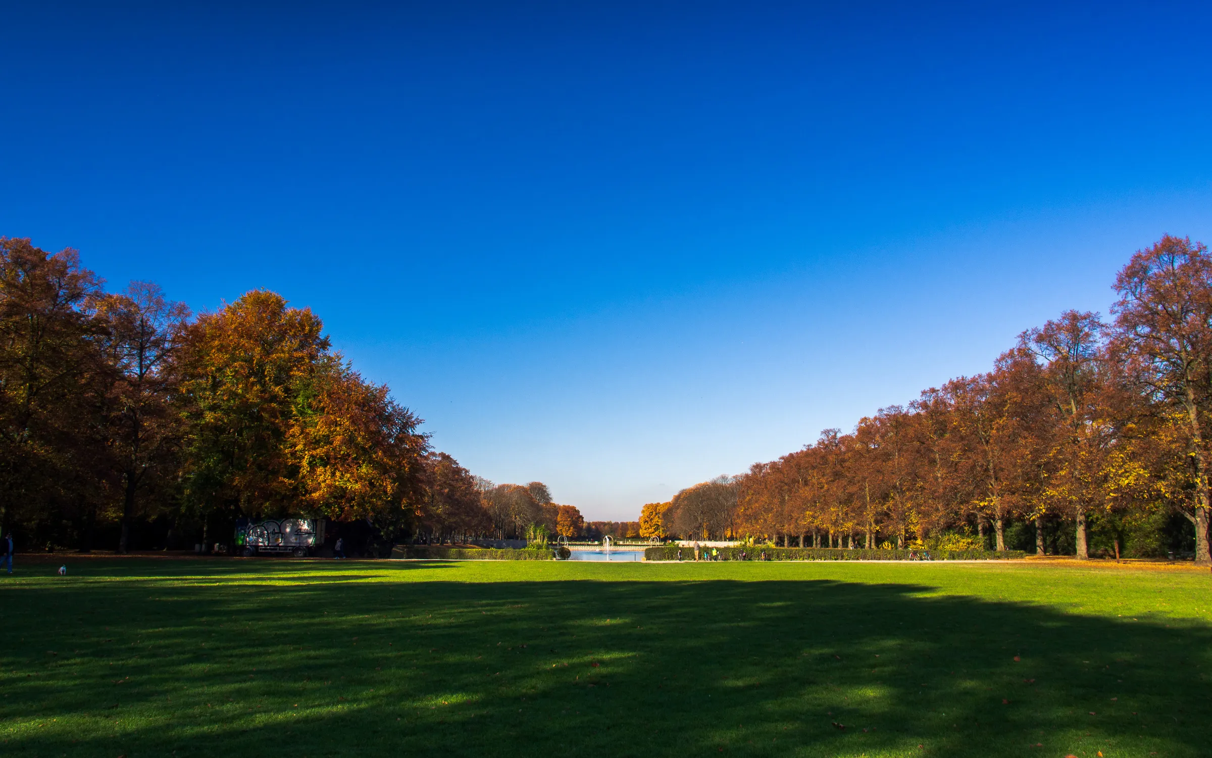 Bright blue sky with scattered white clouds, lush green lawn, trees with autumnal foliage on both sides.