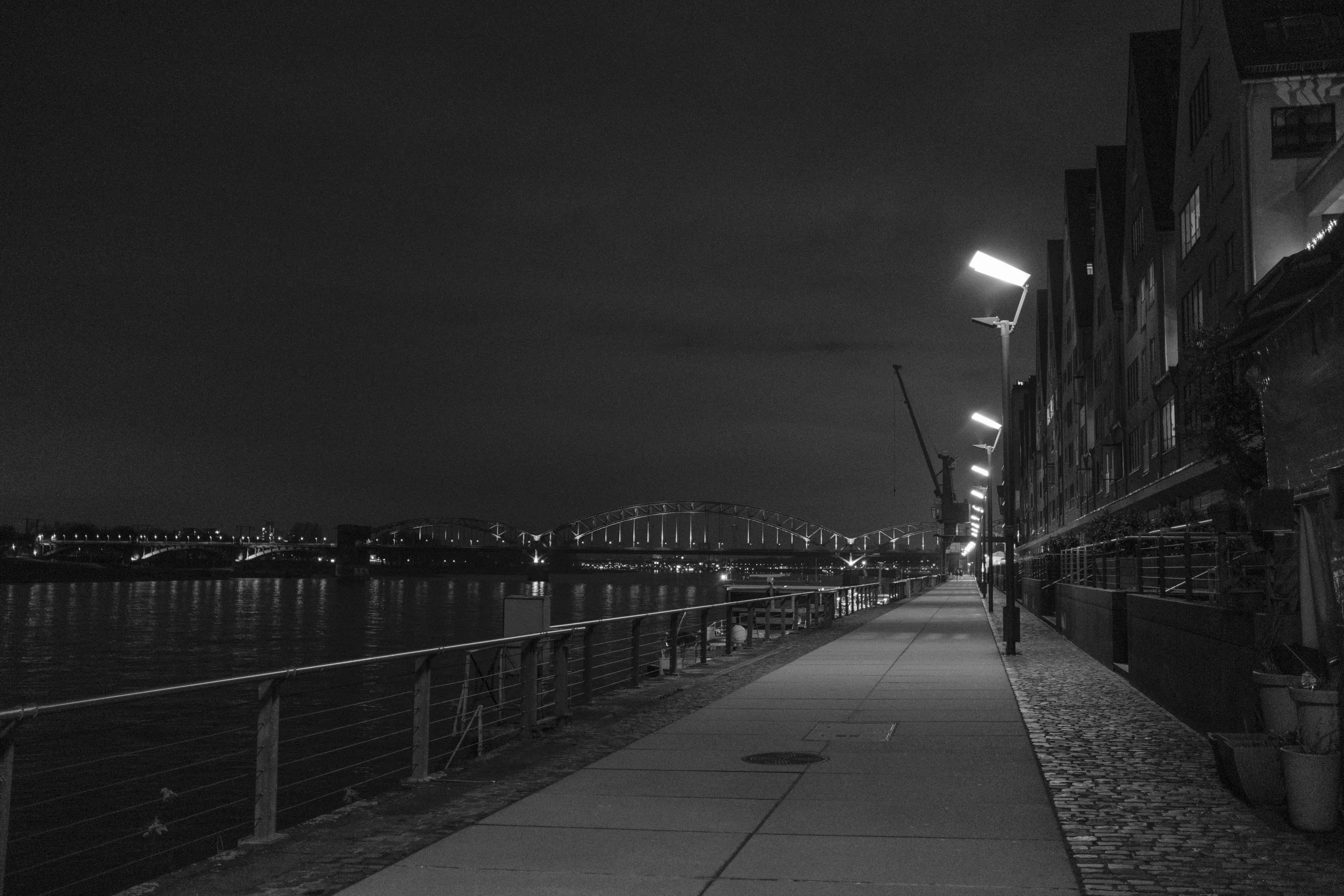 A dimly lit walkway by a river at night, buildings on one side, bridge in distance.