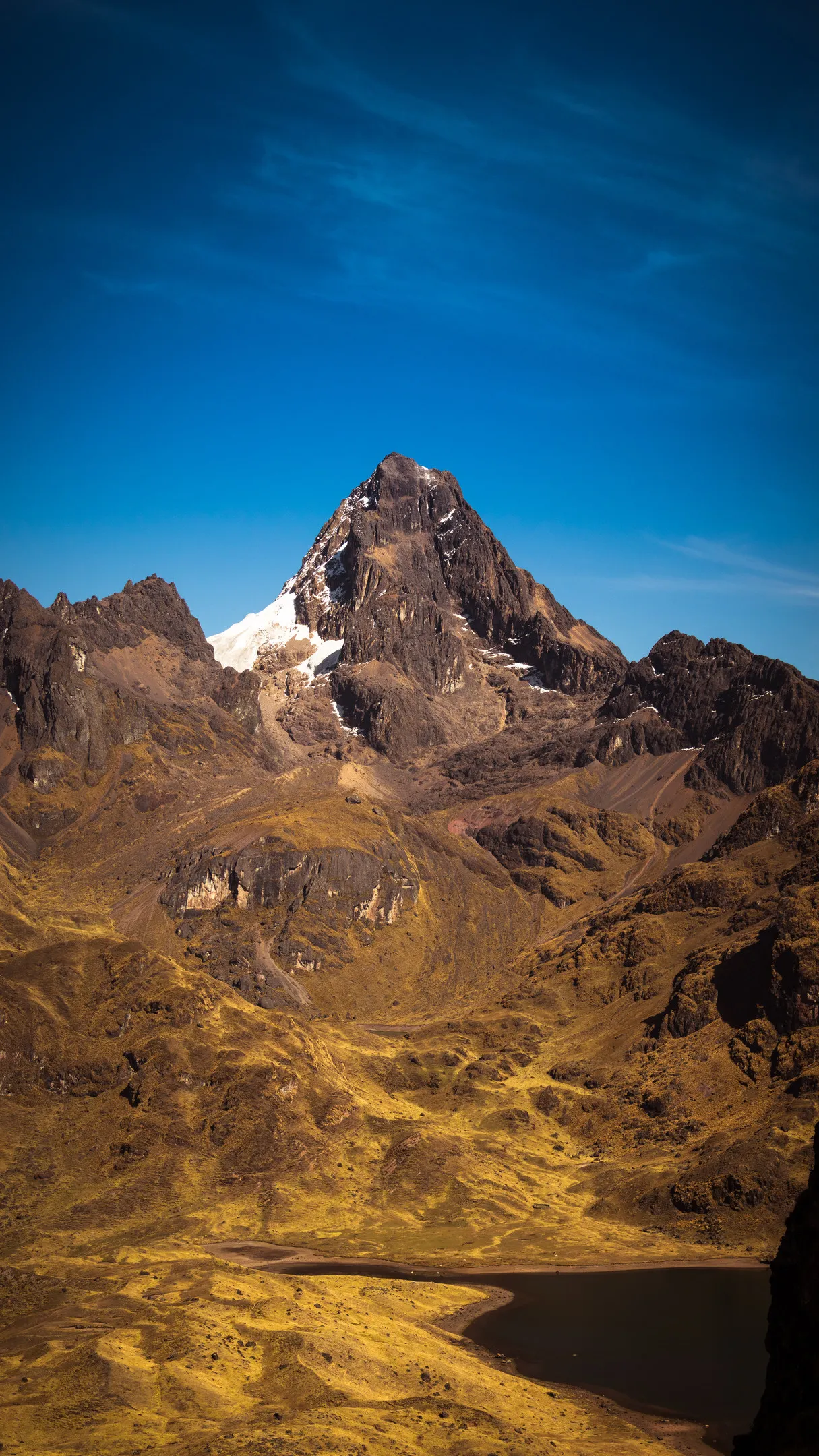 Majestic mountain peak with snow cap, set against clear blue sky, surrounded by barren slopes.