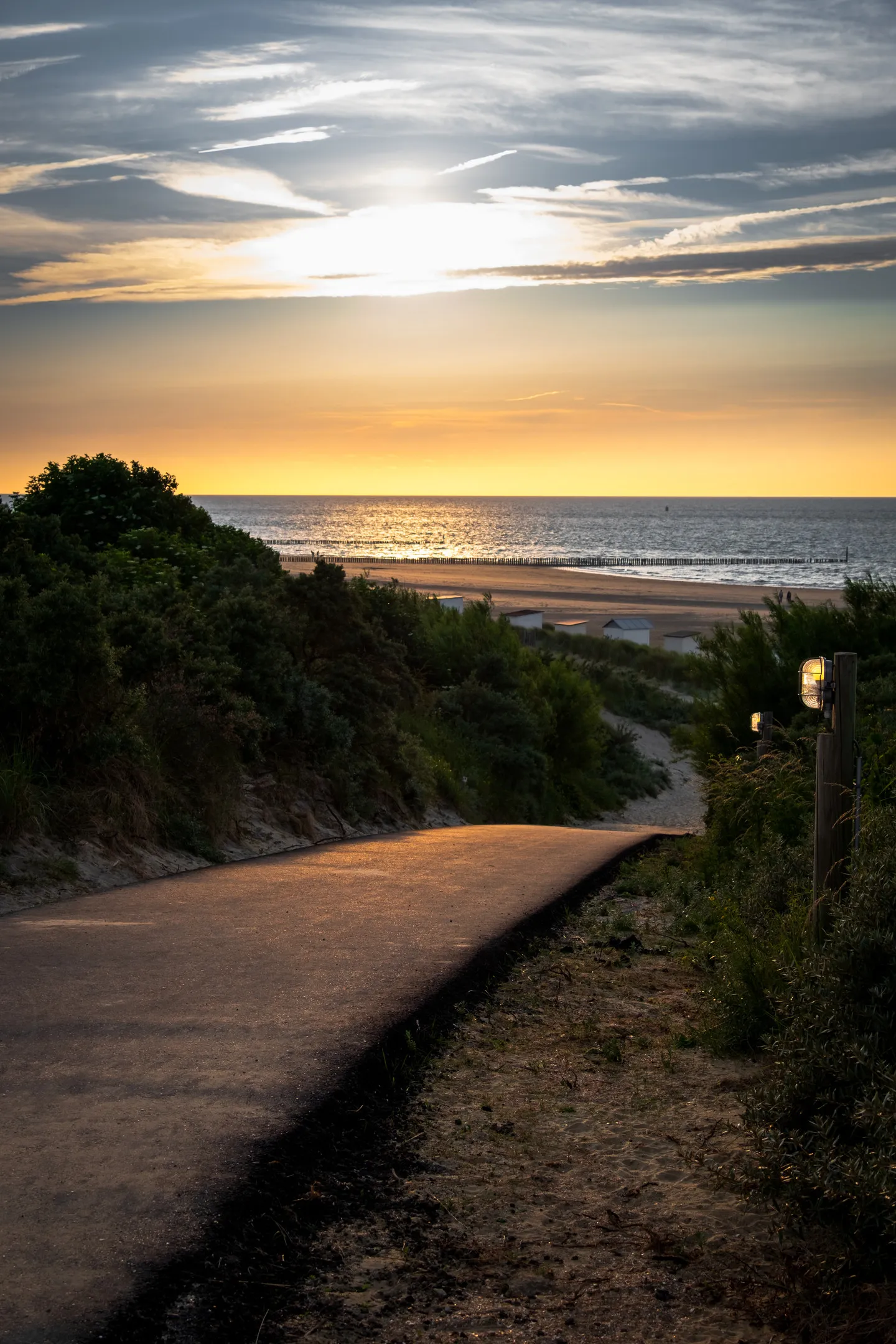 A winding path leads to a beach at sunset with golden light reflecting on the water, clouds scattered in the sky.