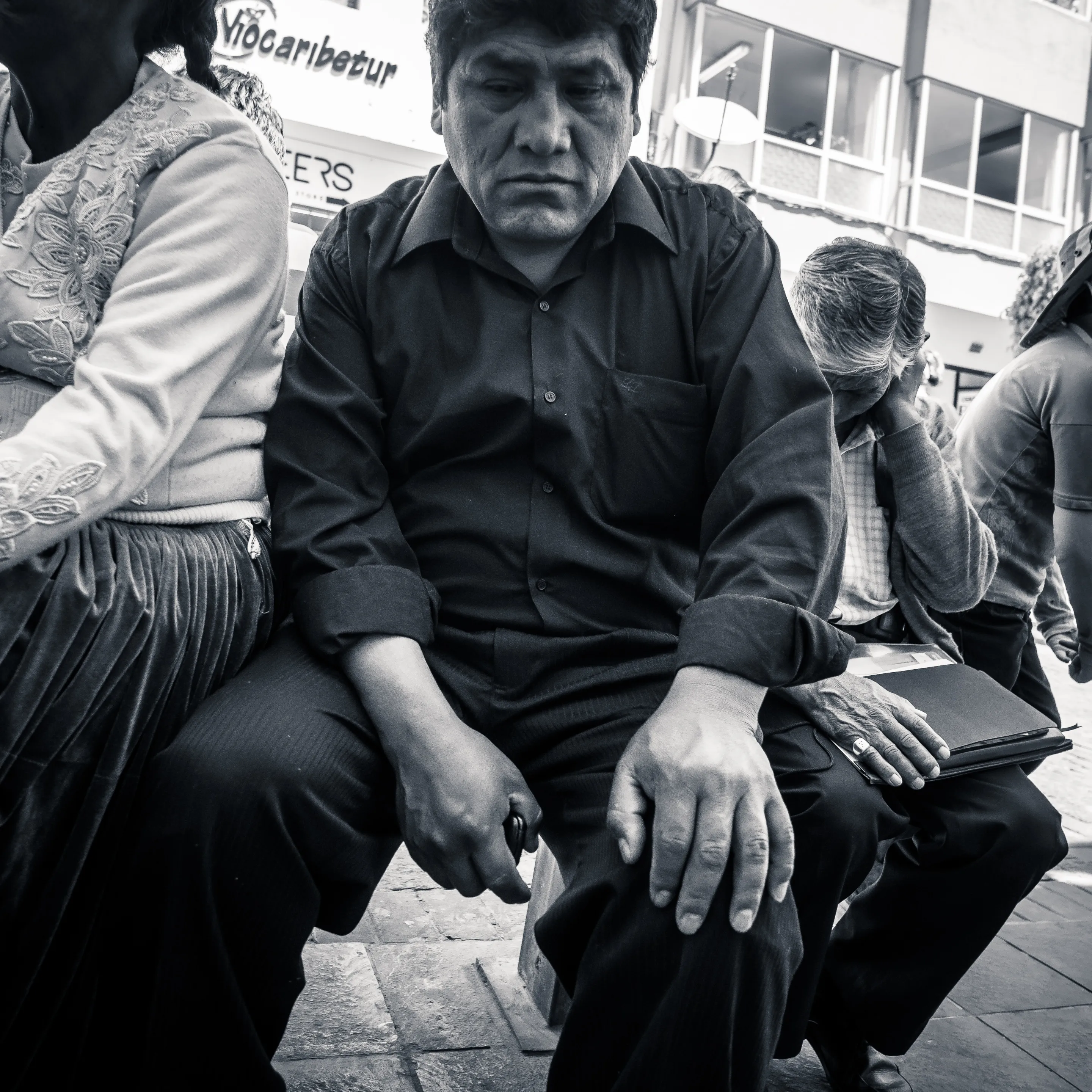 Man in black shirt sitting on bench with hands clasped, surrounded by other seated individuals.