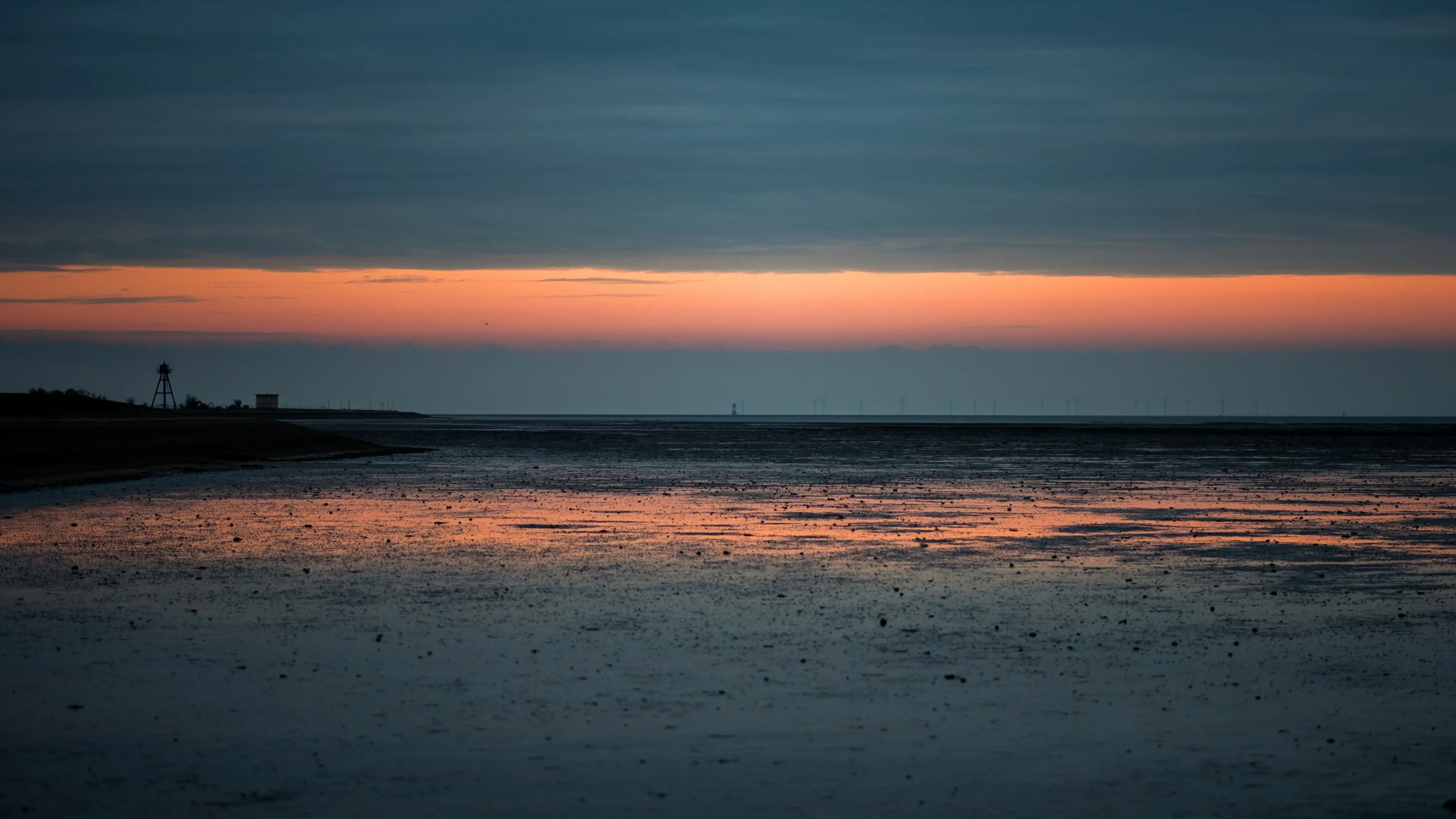 Sunset over calm beach with silhouetted lighthouse in background, vibrant sky reflected on wet sand.