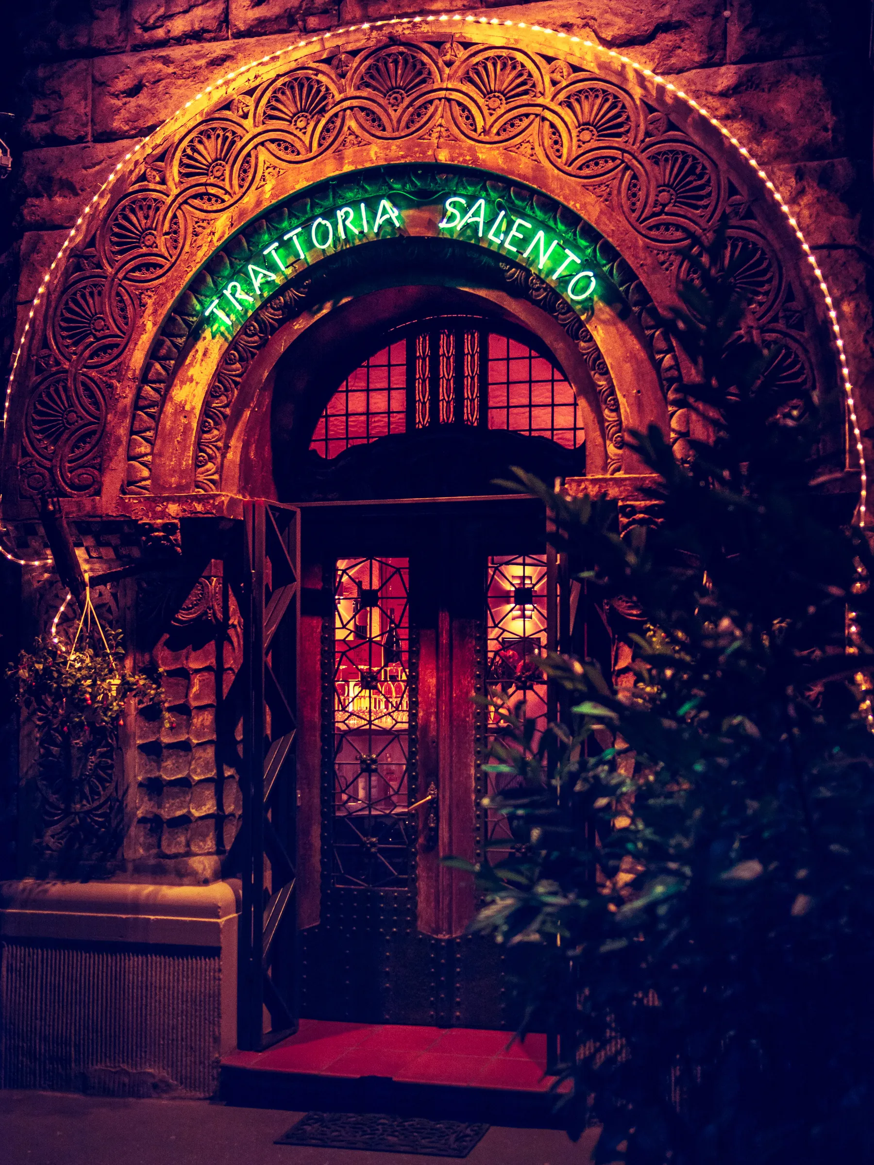 Restaurants entrance with ornate archway, neon sign, plants on either side, red carpet, and glass door.