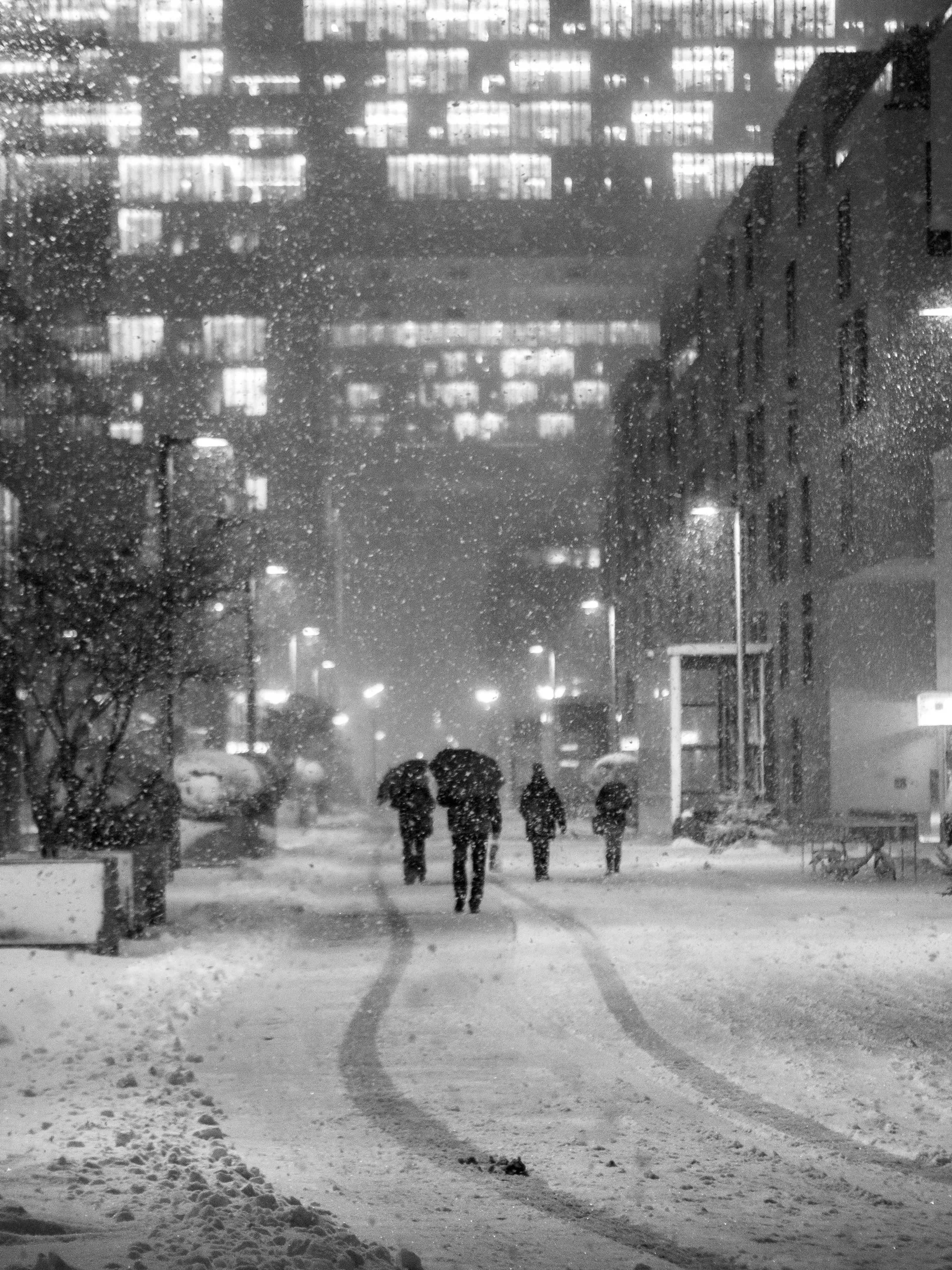 Snowy street at night with people walking, illuminated by warm window lights and street lamps.