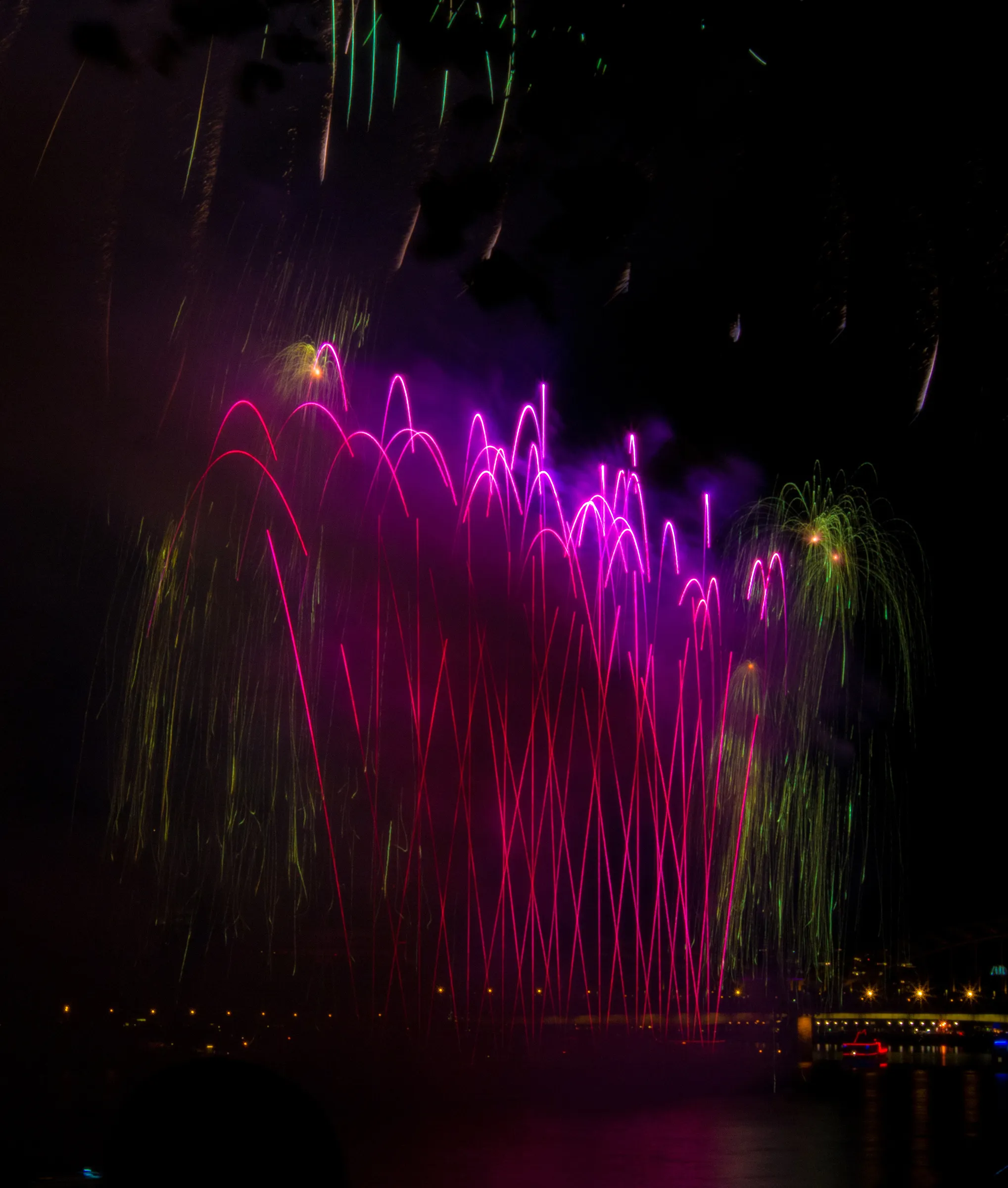 A vibrant fireworks display with pink, purple, and green explosions lighting up the night sky above a body of water.