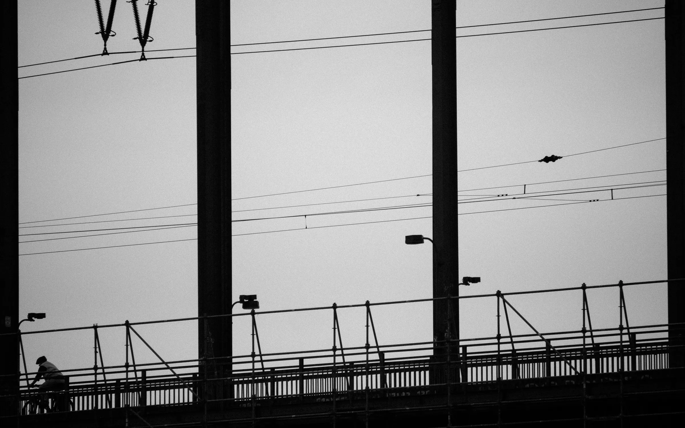 Black and white photo of a bridge with power lines, a bird in flight, and a solitary figure walking.
