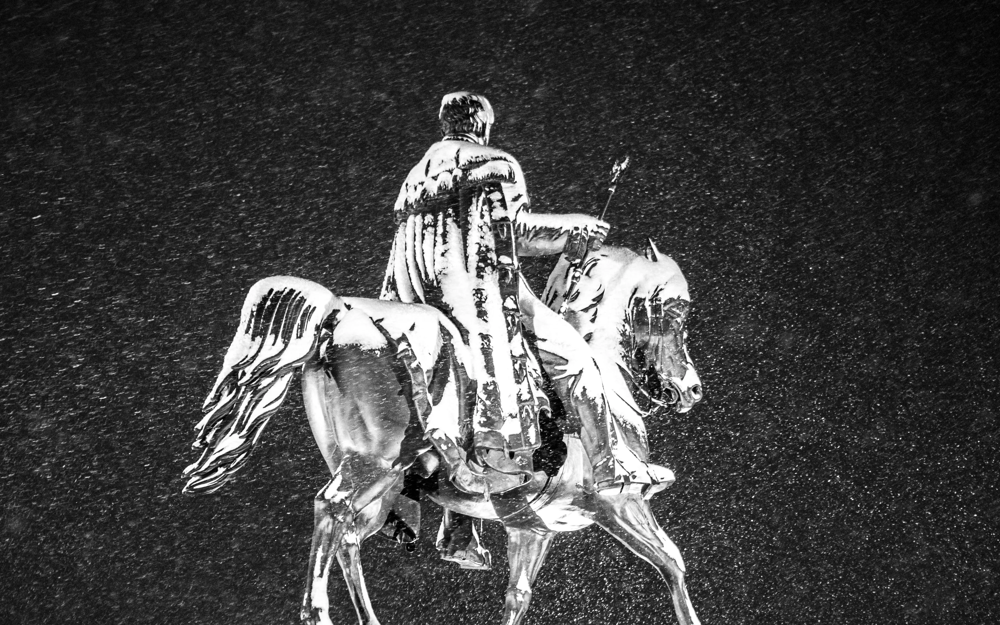 Man in a long coat riding a horse at night, rain falling around them.