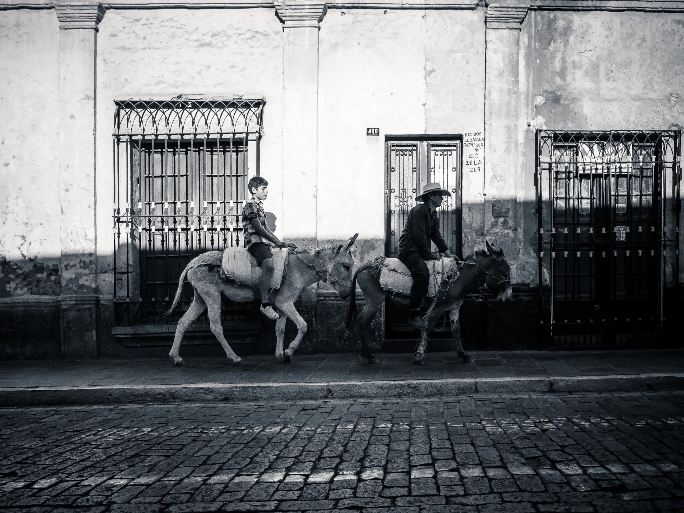 Two people riding donkeys on a cobblestone street, with a weathered building in the background.