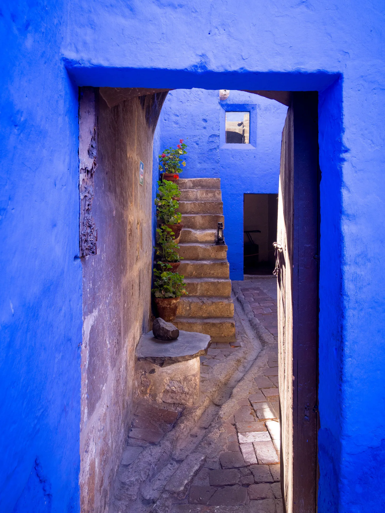 Blue passageway with stone steps, potted plant, and sunlight casting shadows on walls and ground.