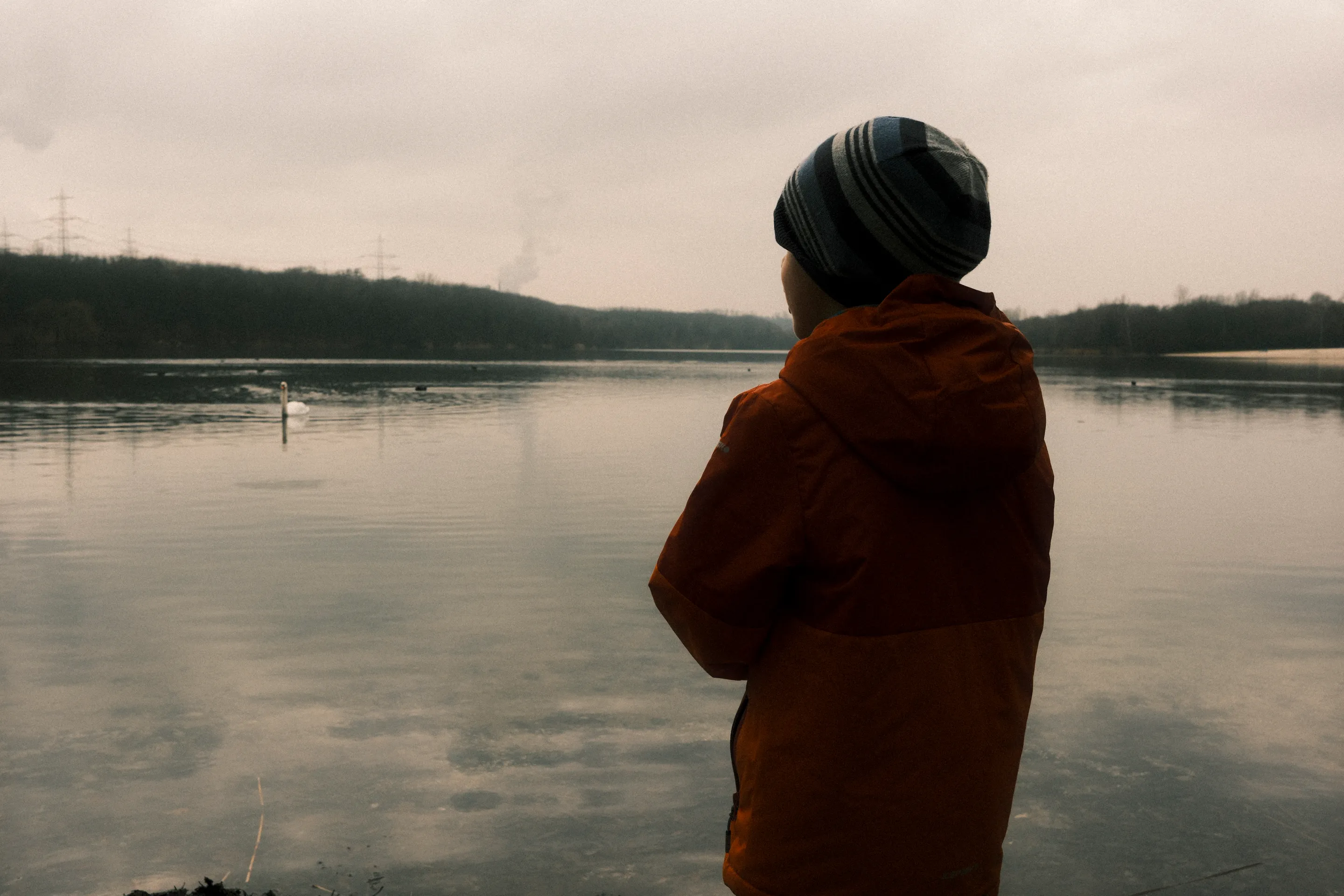Person in orange jacket and gray beanie standing by calm lake, looking away from camera.