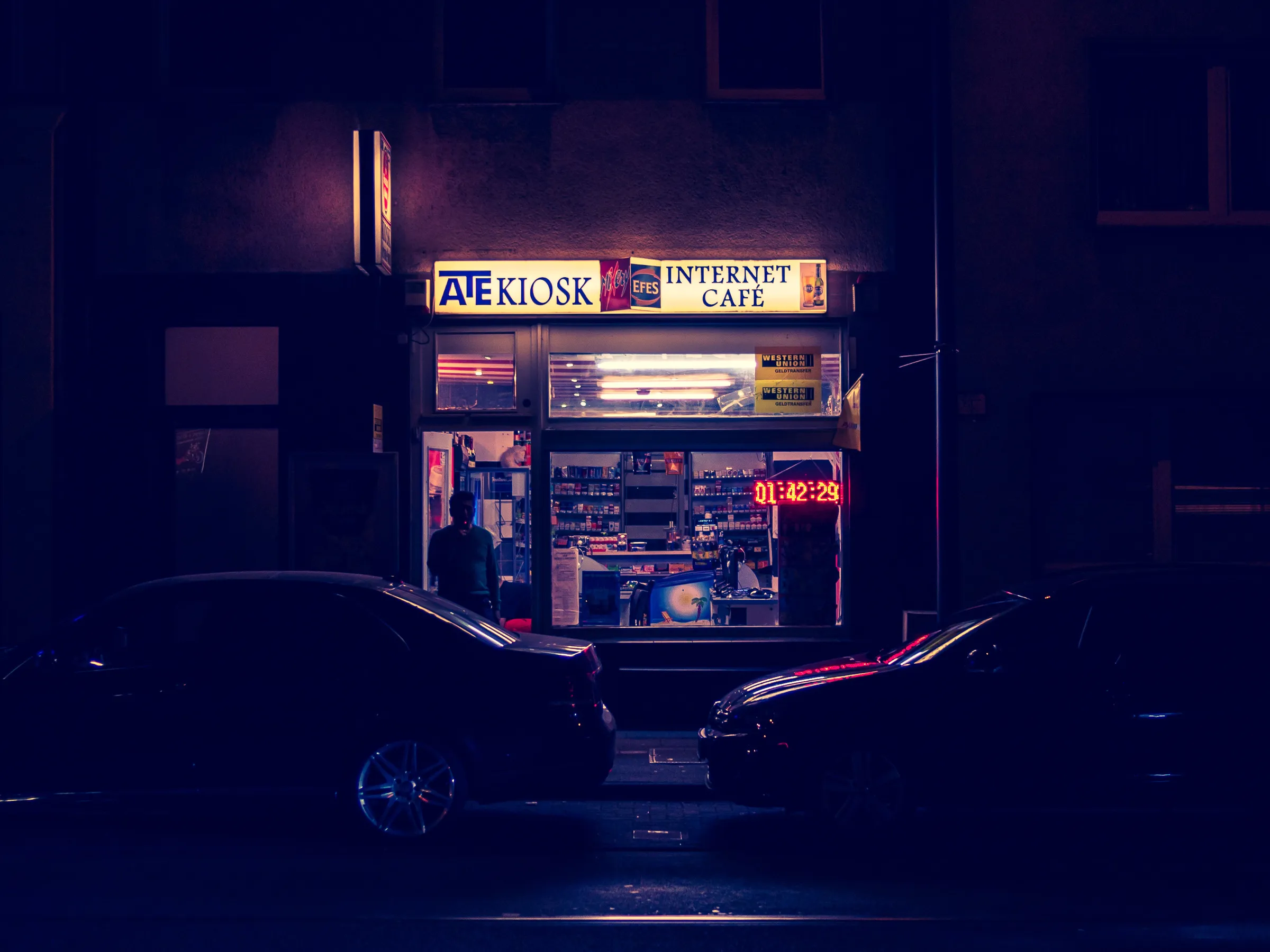 A brightly lit kiosk and internet cafe at night with parked cars in front, displaying snacks and drinks.
