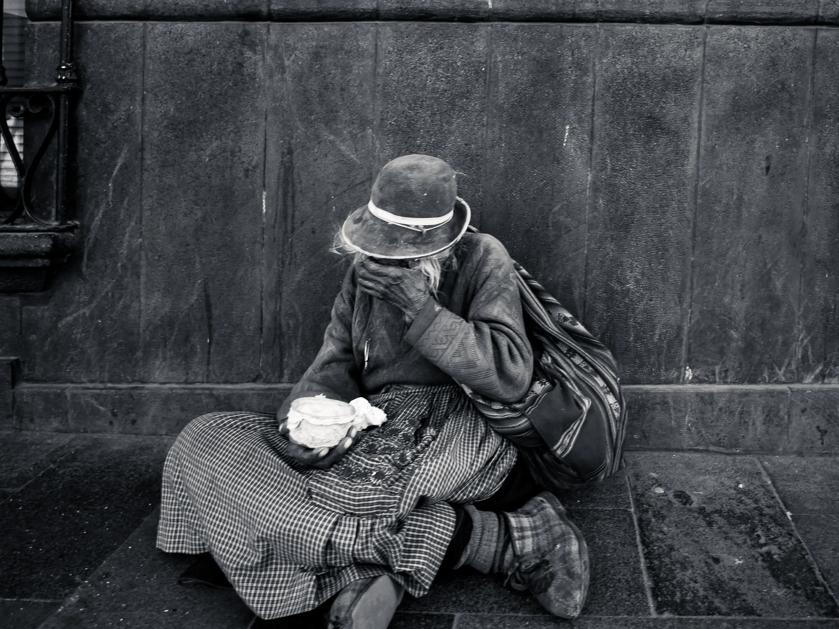 Homeless person sitting against a wall, covering their face with a hat, holding a white object in their lap.