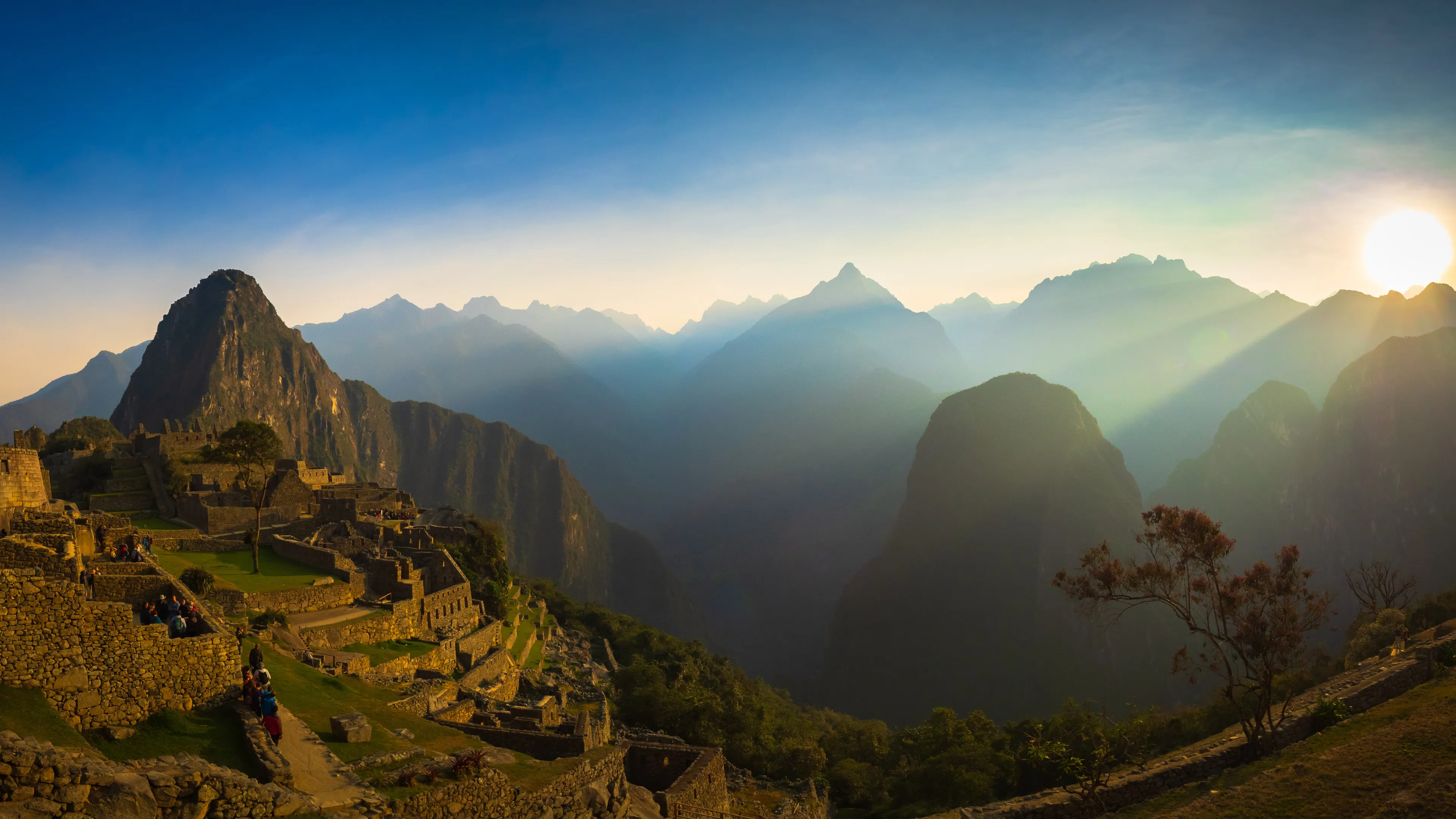 Ancient ruins on a hillside with mountains in the background at sunrise, warm light casting long shadows.