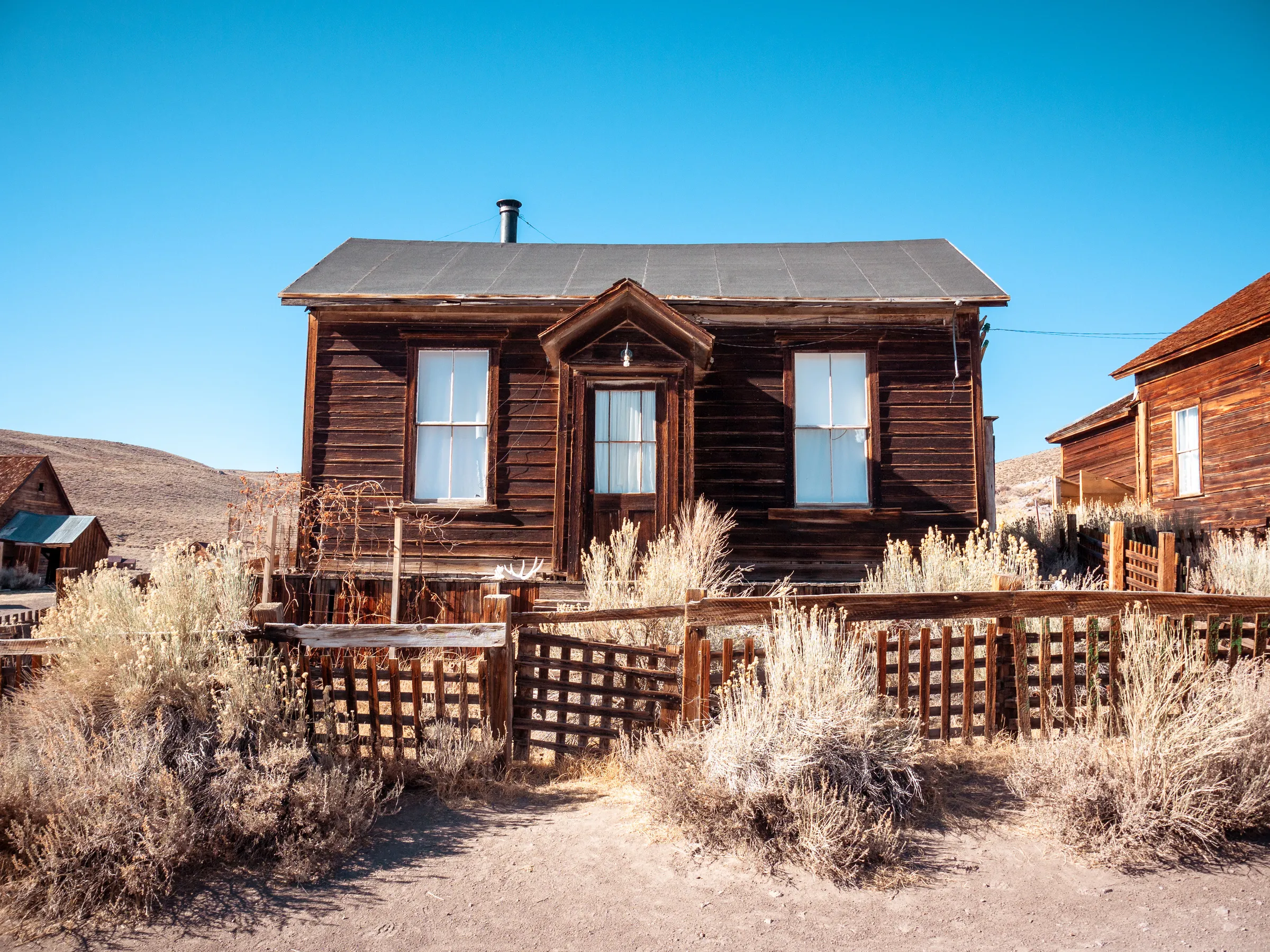 Rustic wooden house with a fenced yard, surrounded by dry grass and desert landscape.
