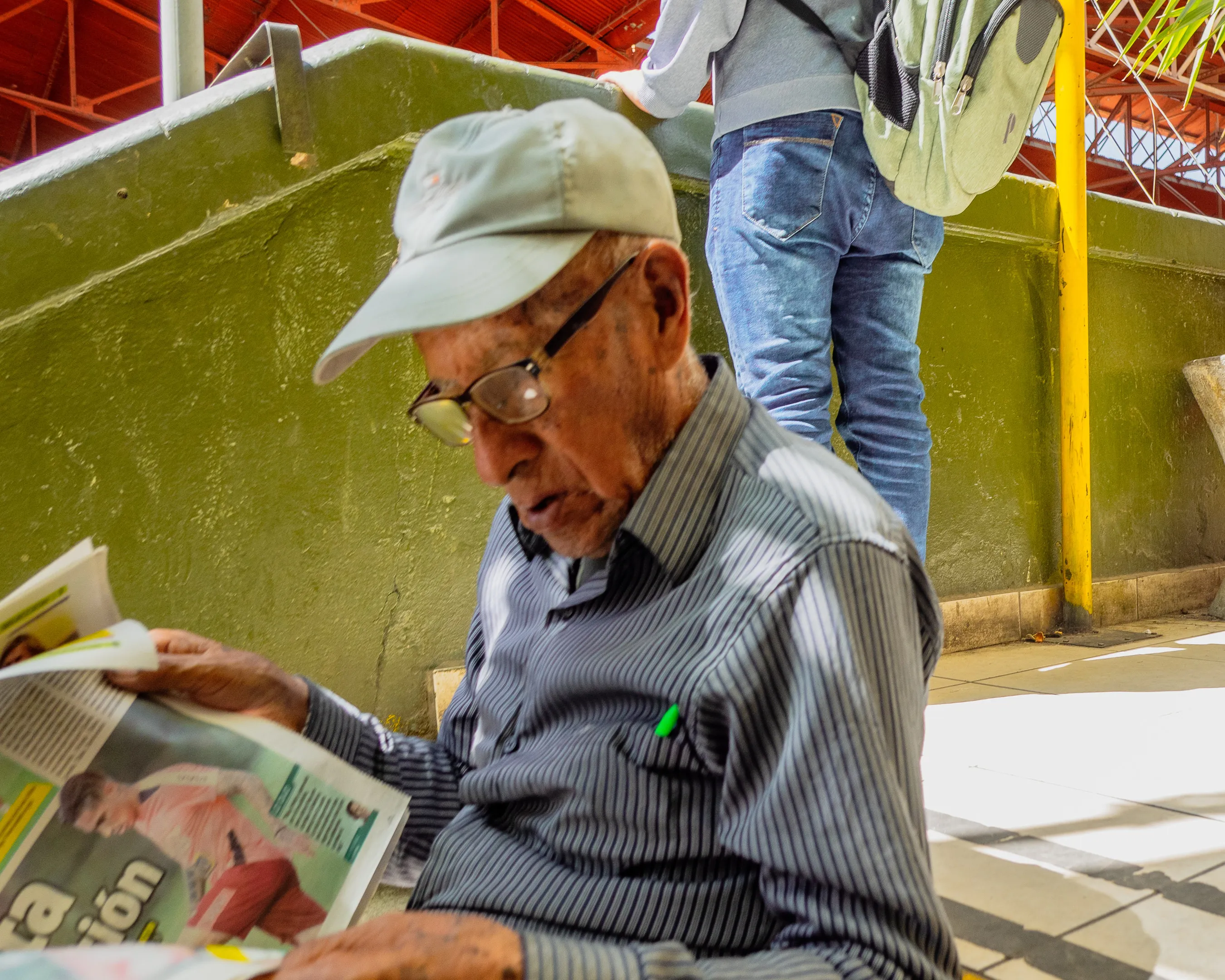 Elderly man in cap reading magazine on a bench, people walking past, green wall and yellow railing behind.