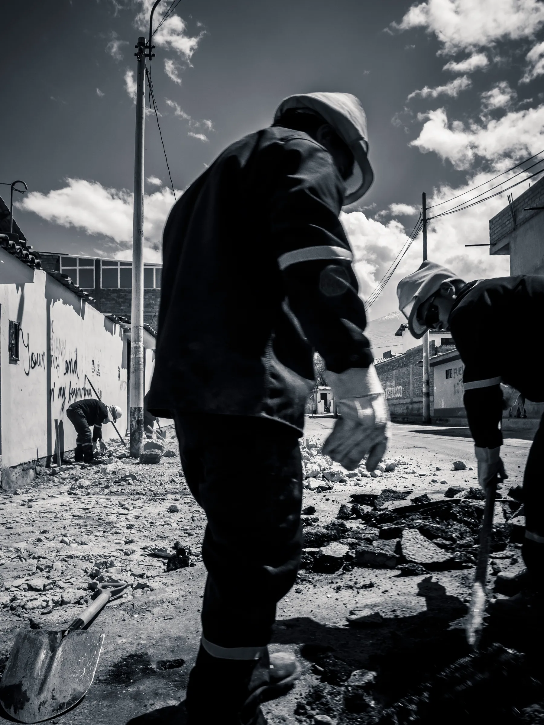 Three workers in helmets, two standing, one digging, debris on street, urban setting, black and white photo.