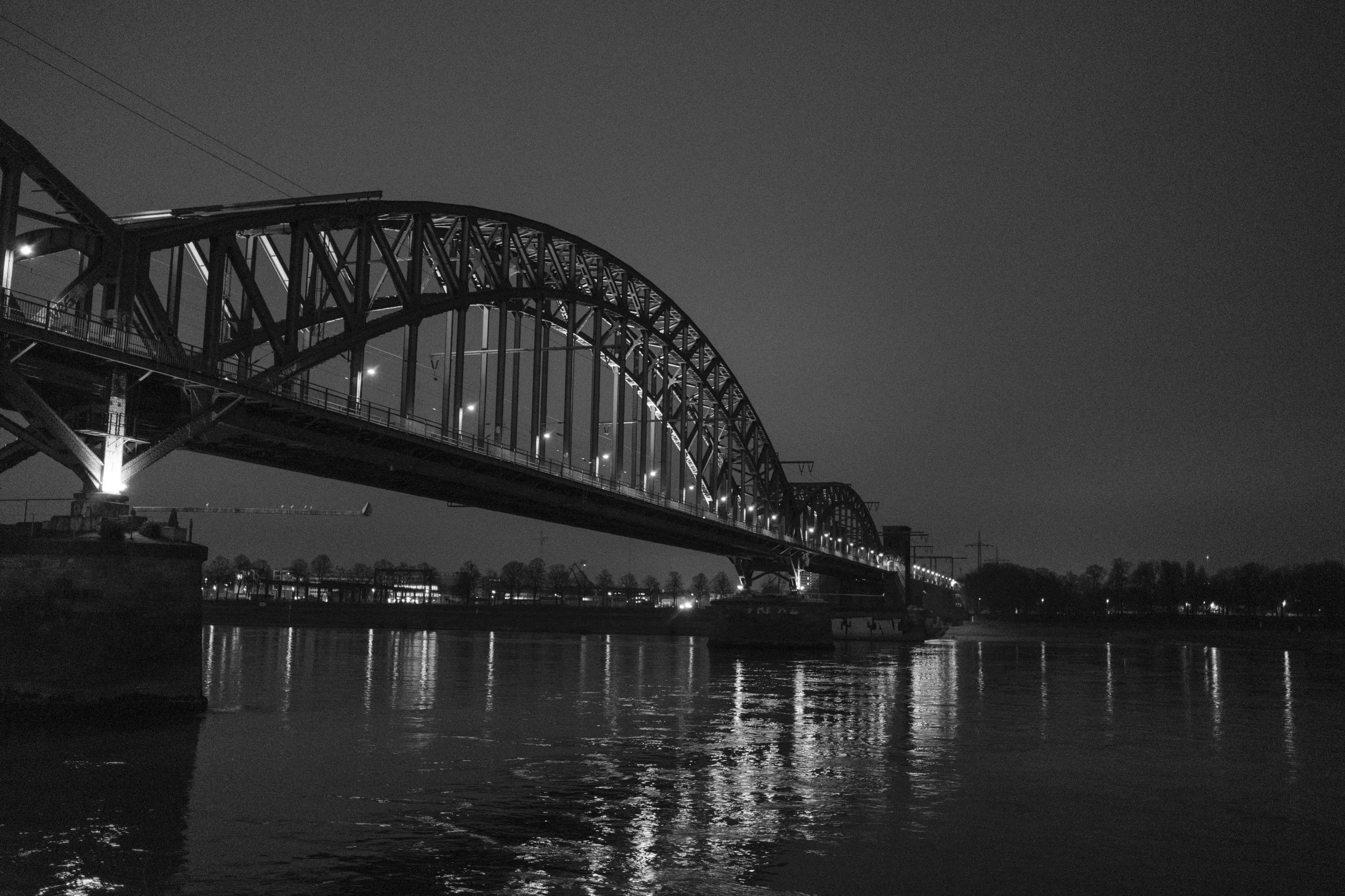 Illuminated bridge over calm river at night, silhouetted against dark sky.