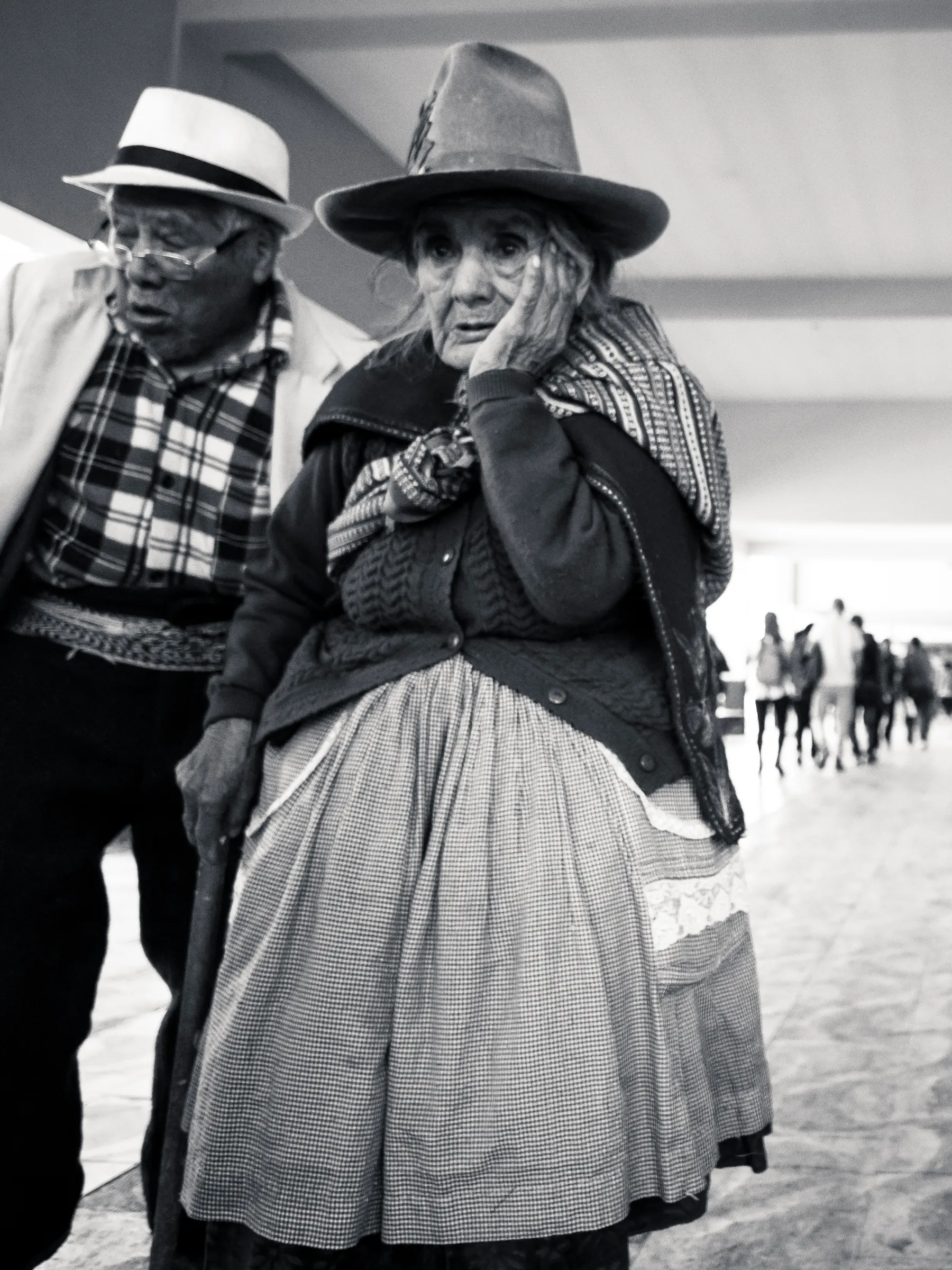Woman in hat and plaid shawl, hand on face, man in hat and checkered scarf, blurred figures in background.