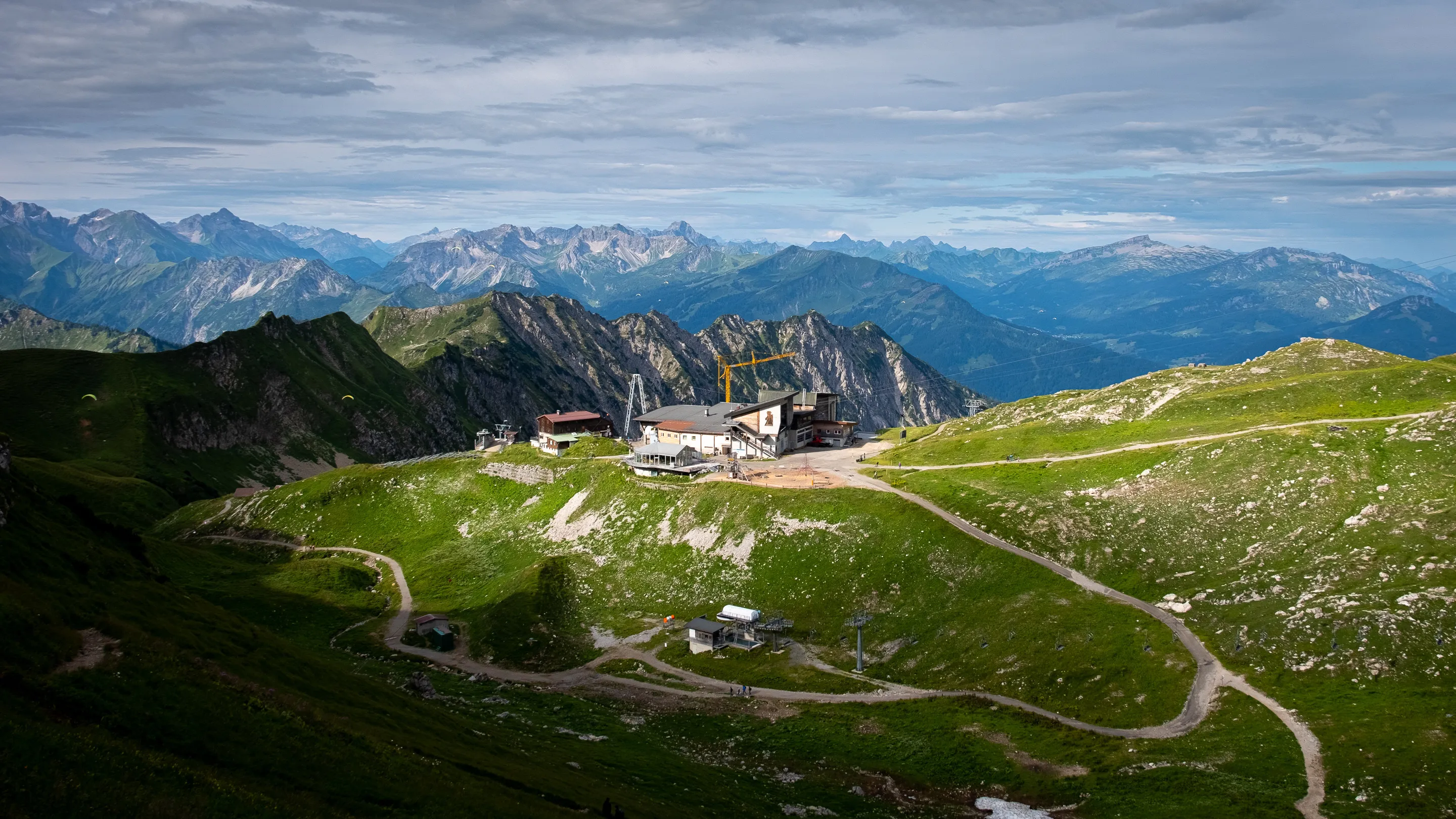 Mountain landscape with a building near a winding road, surrounded by rolling green hills and distant peaks under cloudy sky.