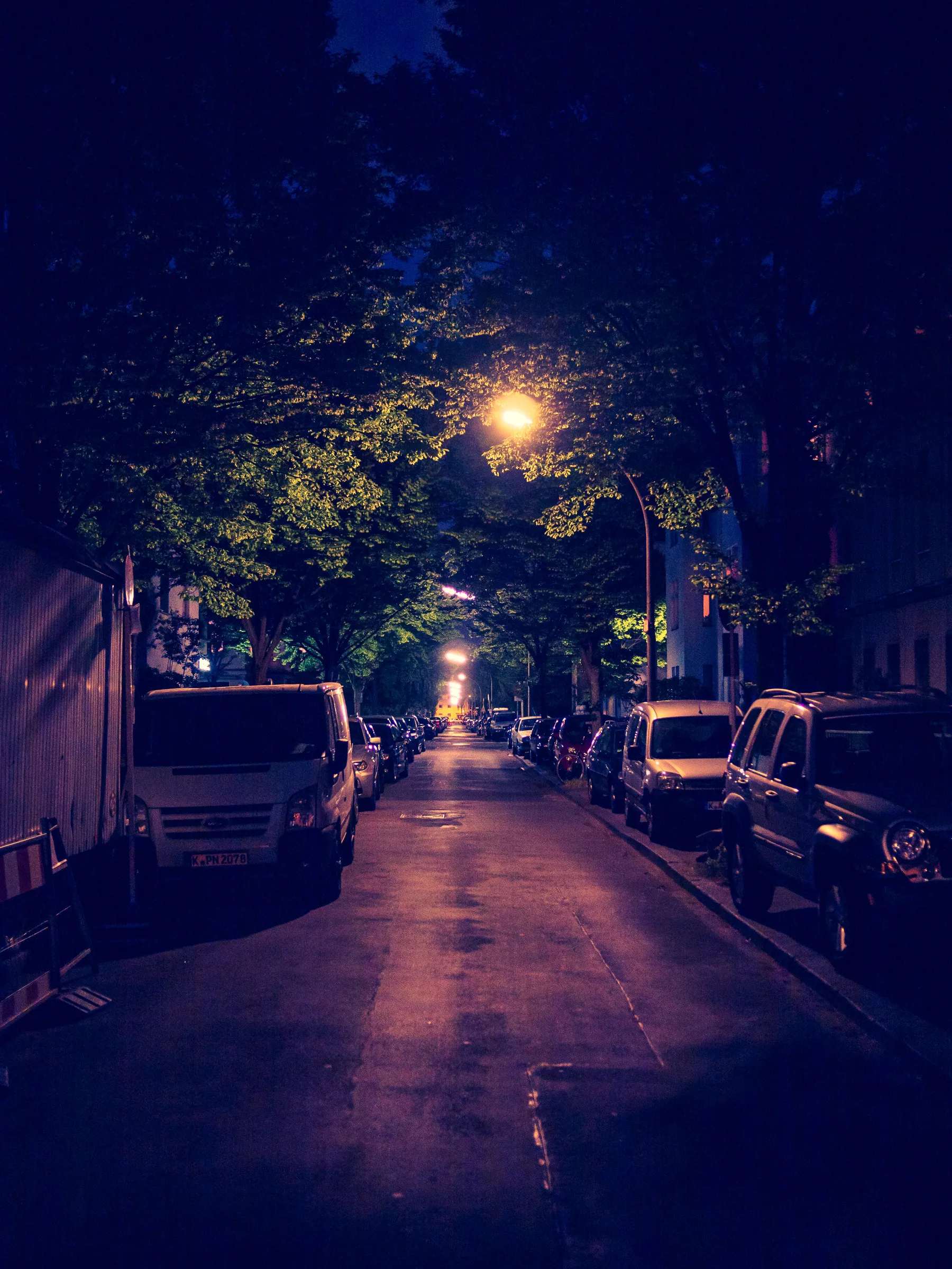 Dimly lit street at night, lined with parked cars and trees, a single lamp post glowing in the distance.