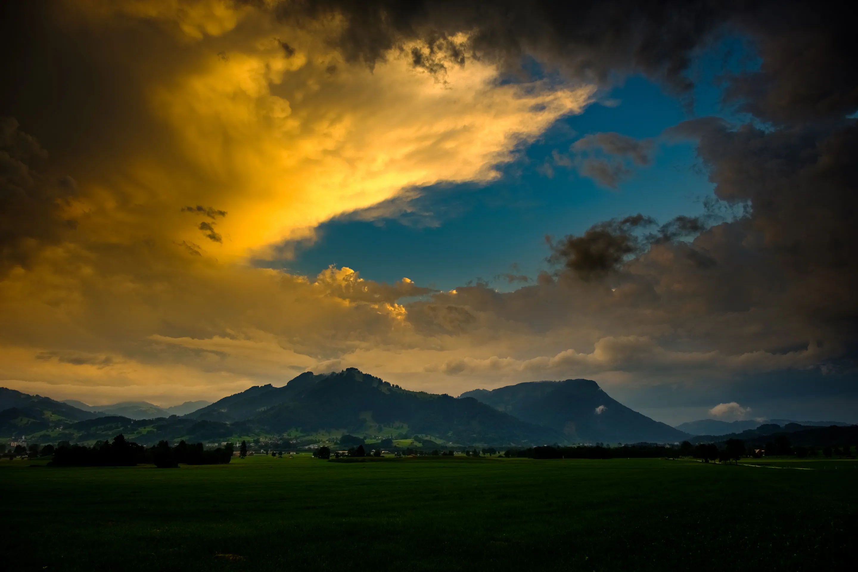 Golden sunset light breaking through dark clouds over green field with distant mountains.