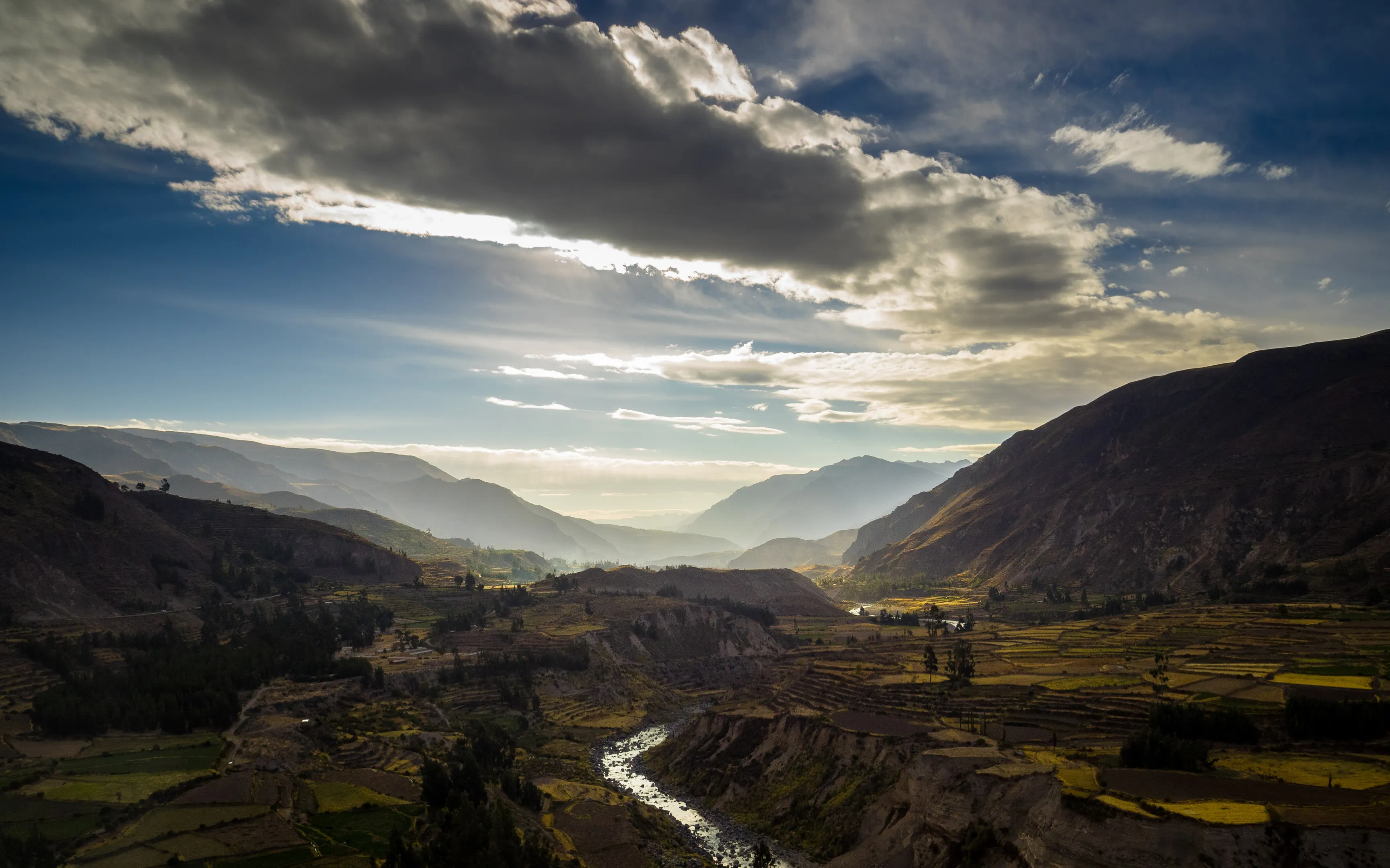 Mountainous landscape with a river winding through terraced fields beneath dramatic clouds at sunset.