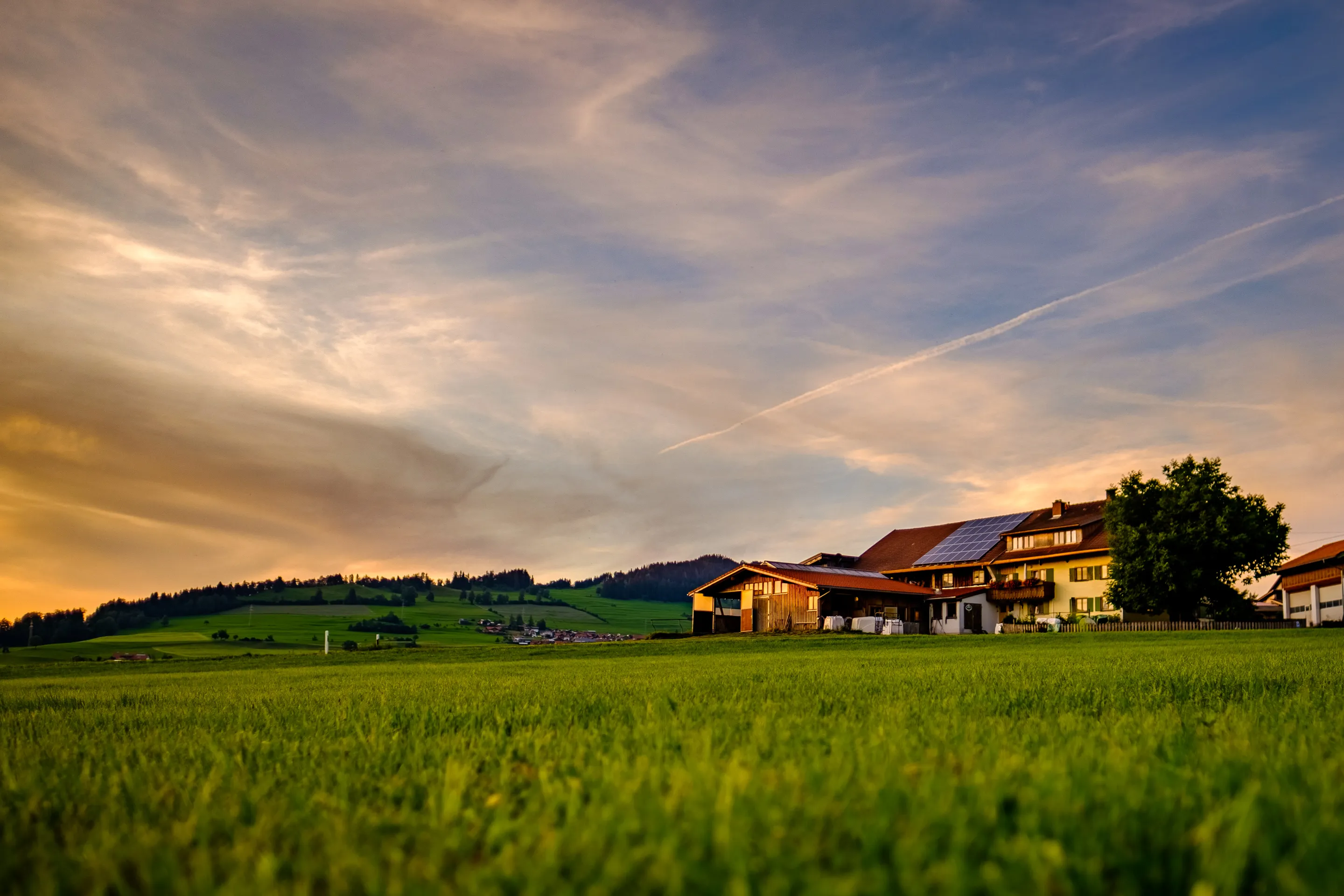 Golden light on farmhouse in lush green field with dramatic sunset sky and distant hills.