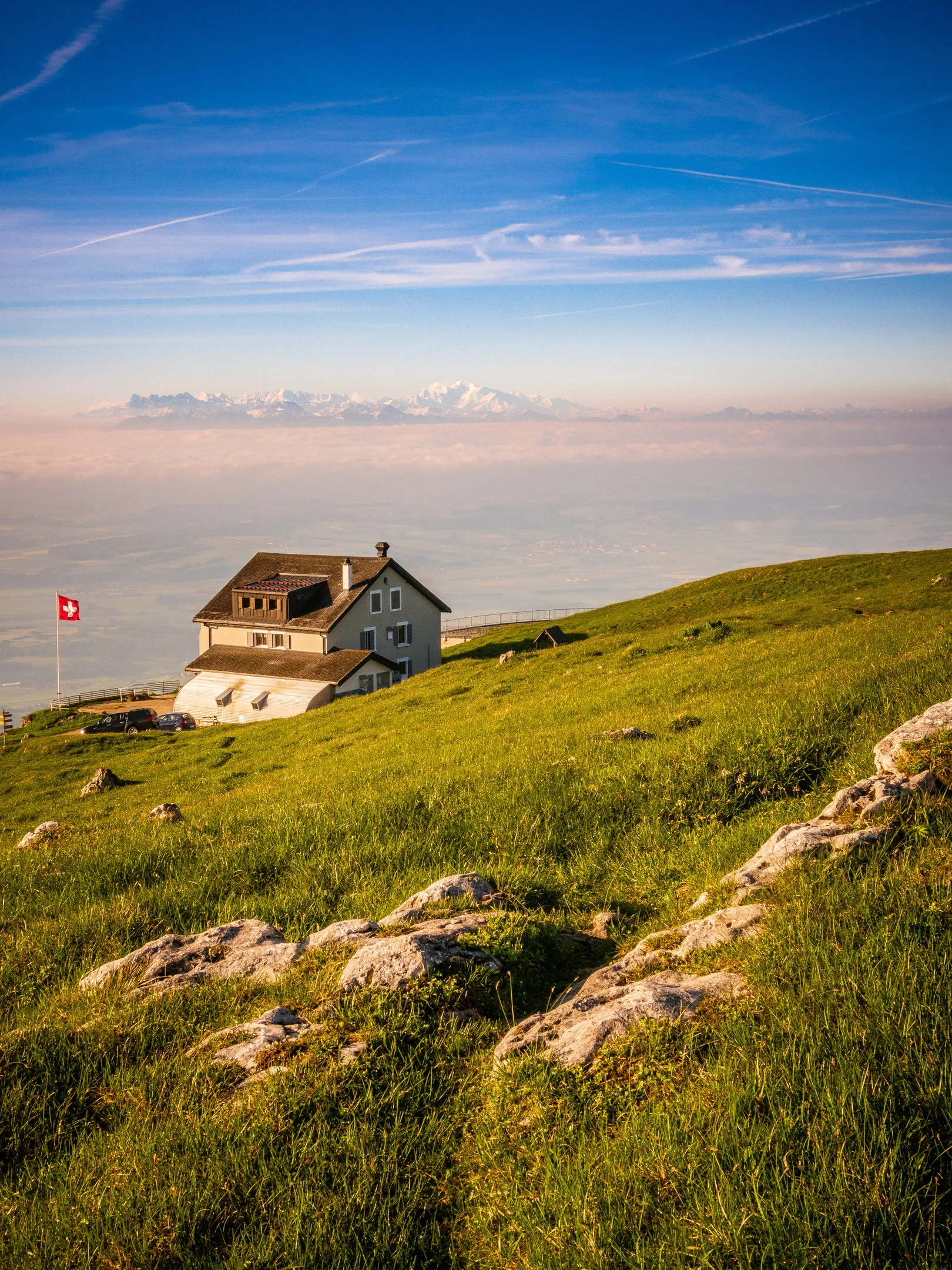 White house on green hillside, Swiss flag flying, mountains in background under blue sky with clouds.