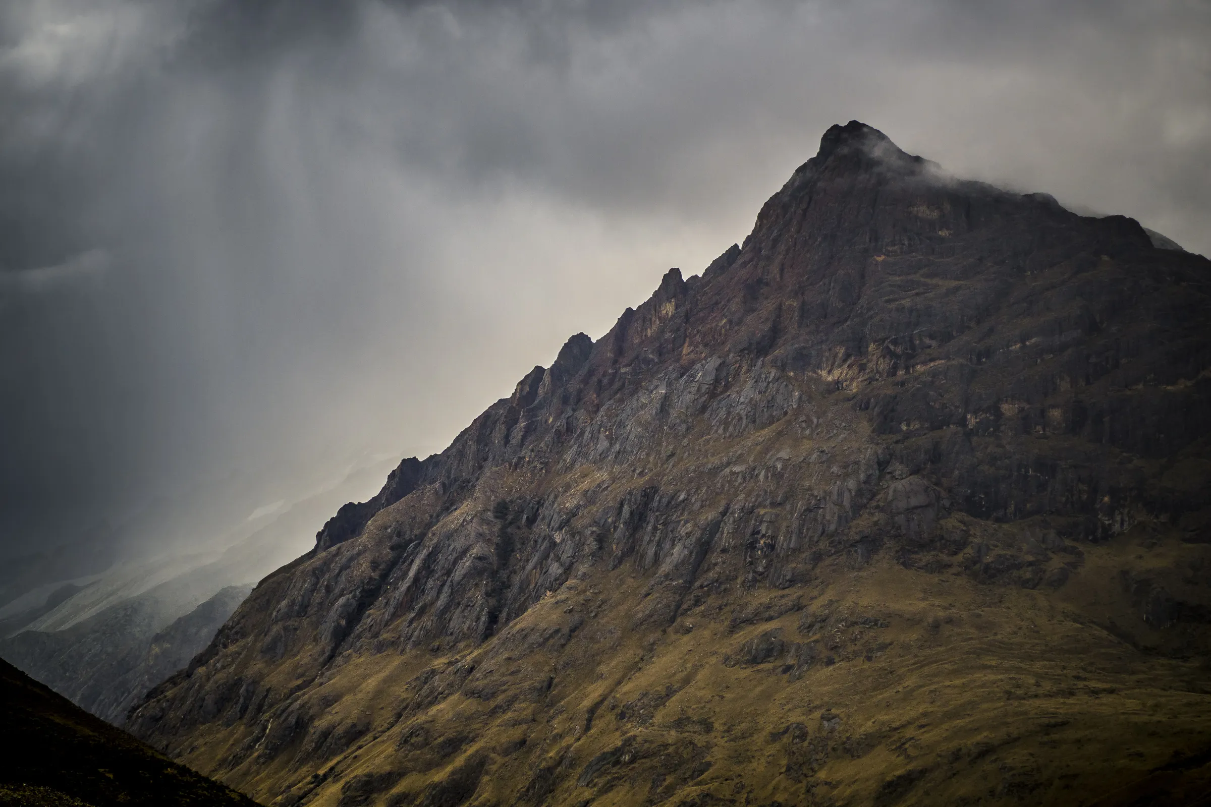 Dark, moody mountain peak with rocky face, set against dramatic cloud-filled sky.