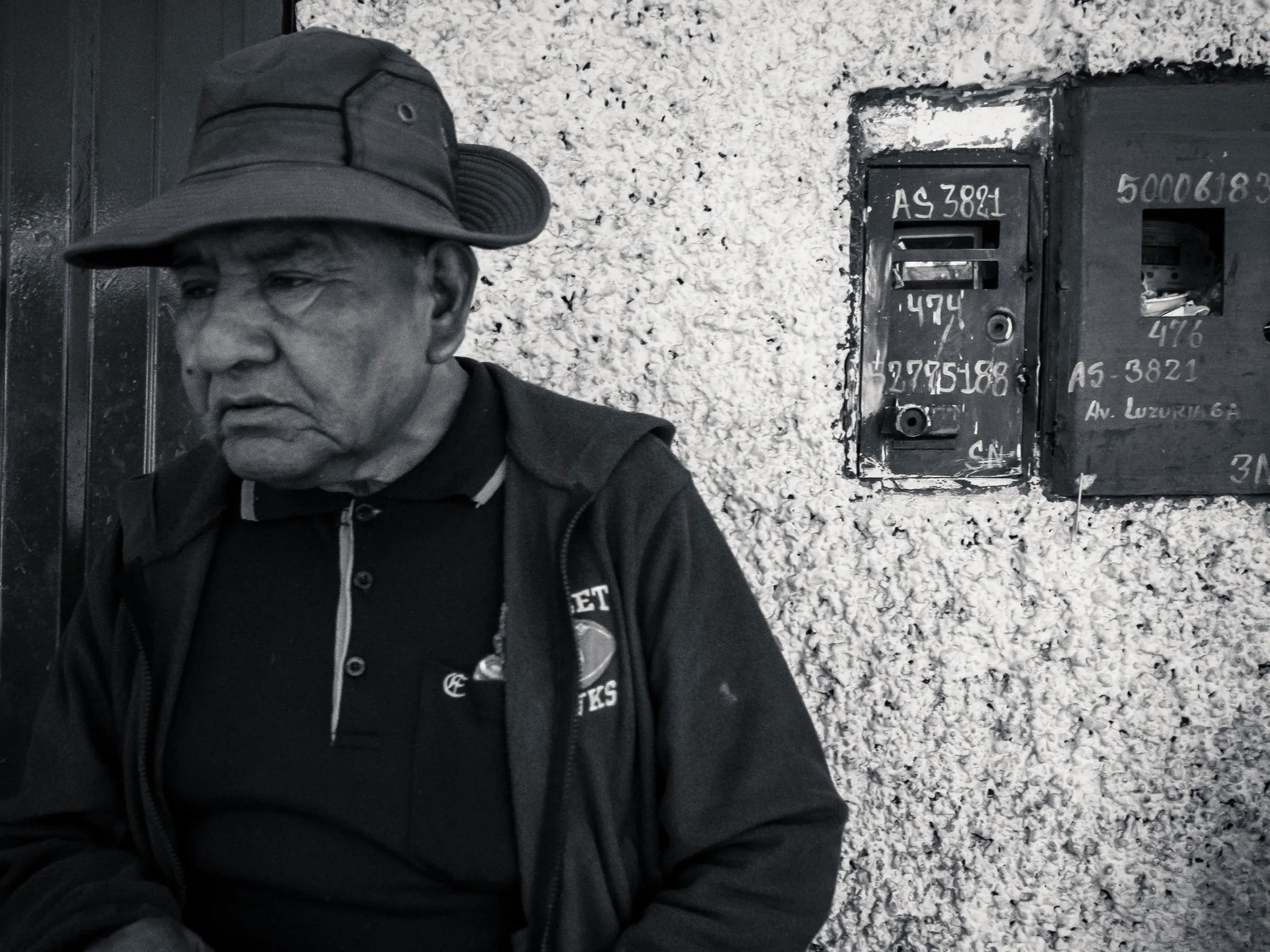 Old man in hat sitting against wall with mailboxes behind him, black and white photo.