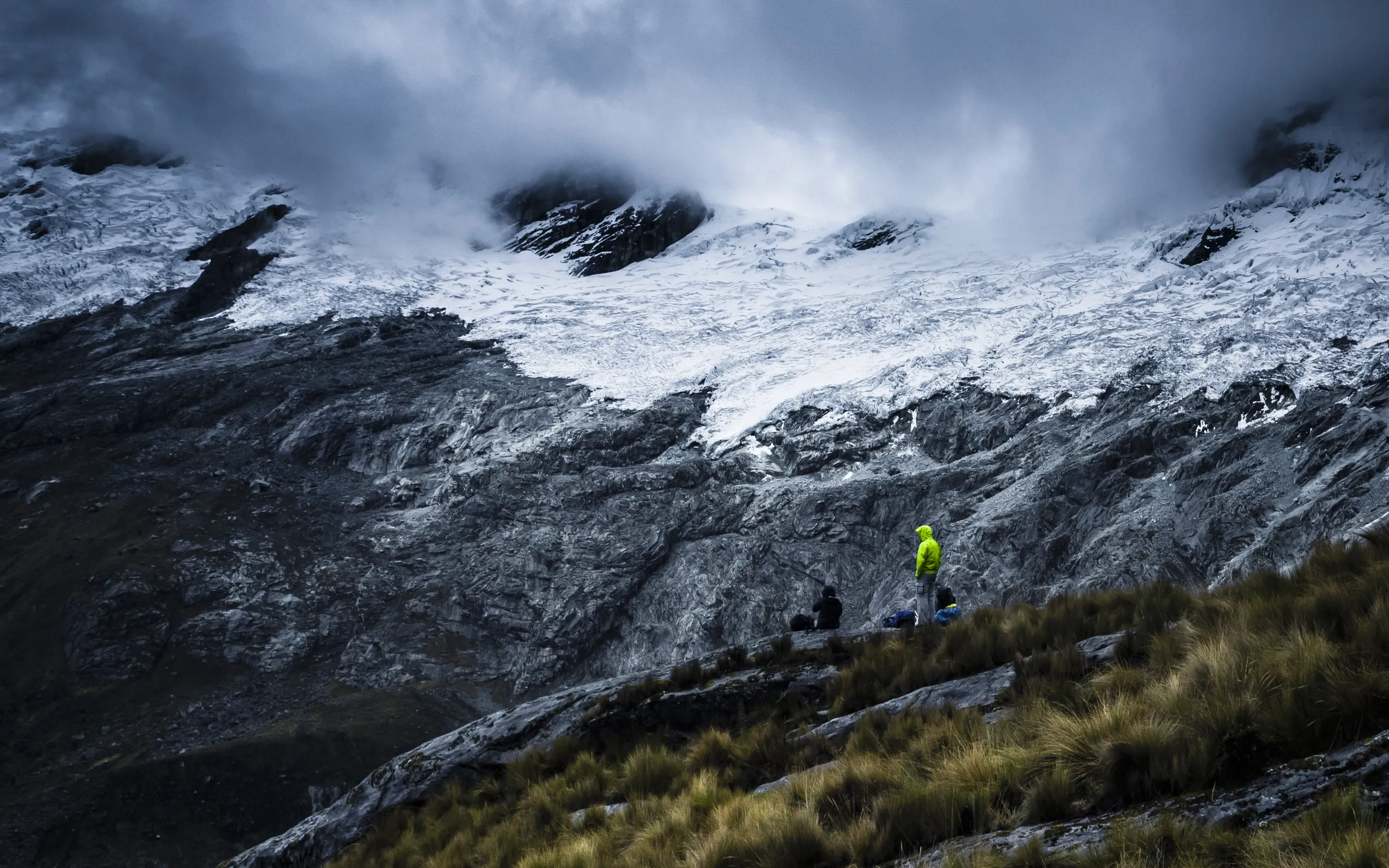 Two hikers on a rocky trail with snow-covered mountains in the background and low clouds.