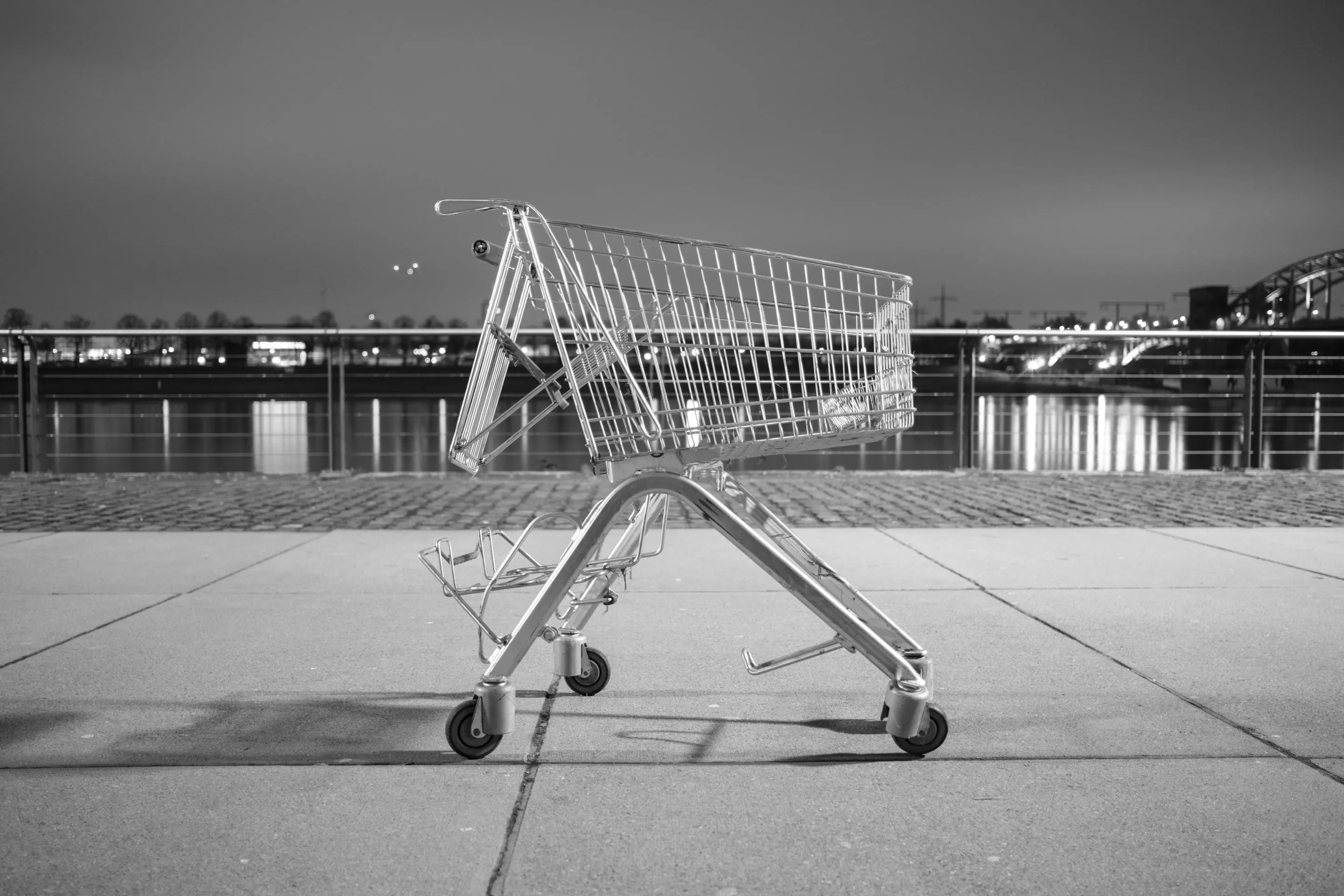 Empty shopping cart tipped on its side, wheels facing upward against an urban waterfront at night.