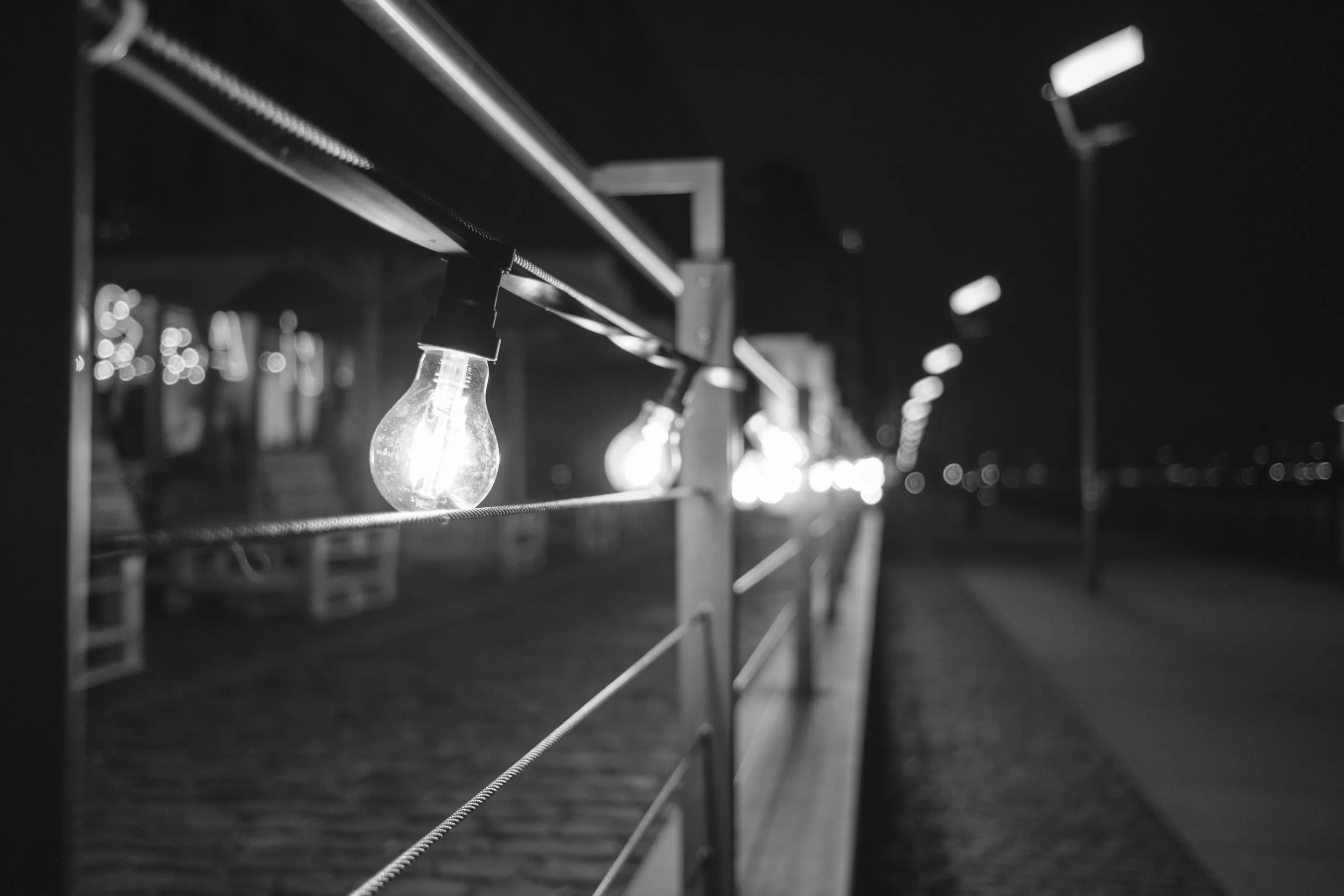A glowing lightbulb hanging from a wire in a nighttime setting with blurred lights in the background.