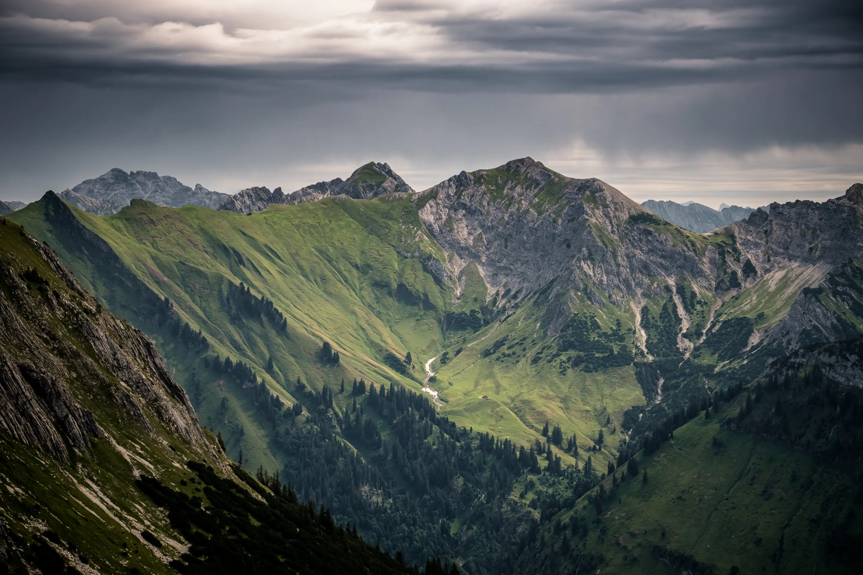 Mountainous landscape with green valleys and forests under cloudy sky.