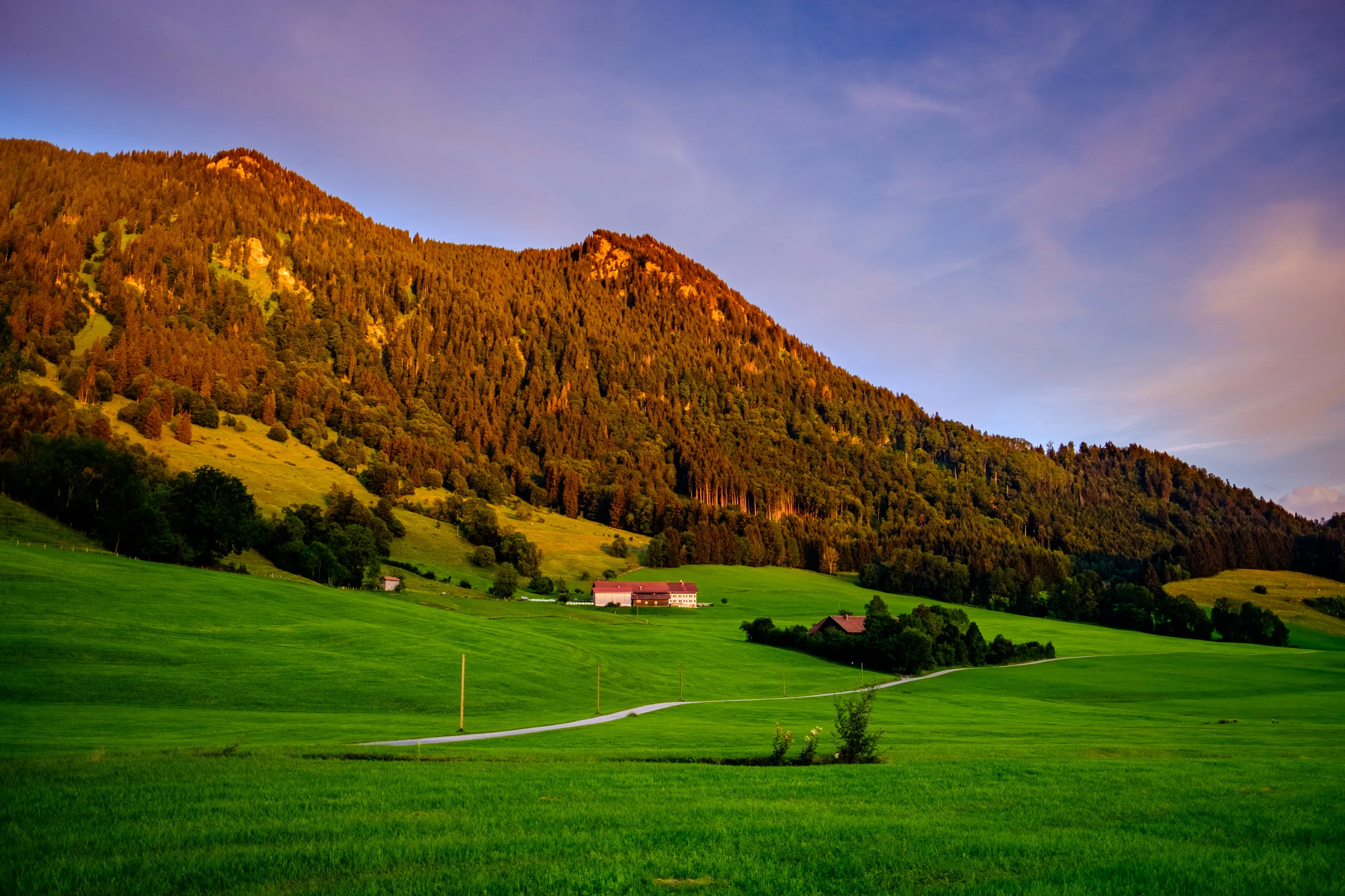 A mountain at sunset with a farmhouse in the foreground, green fields and a winding path.