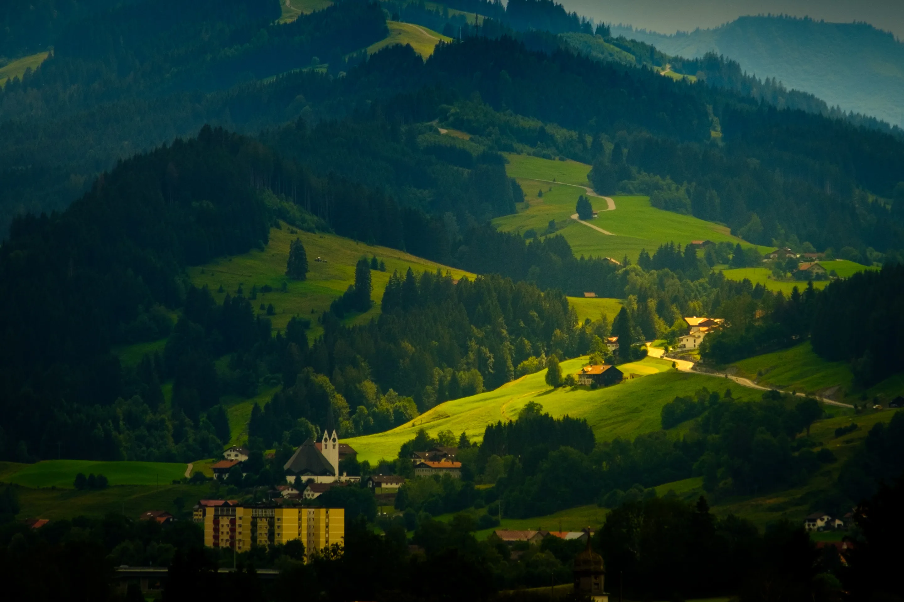 Rolled hills with a church in the foreground, lush green fields and trees, hazy mountains in the distance.