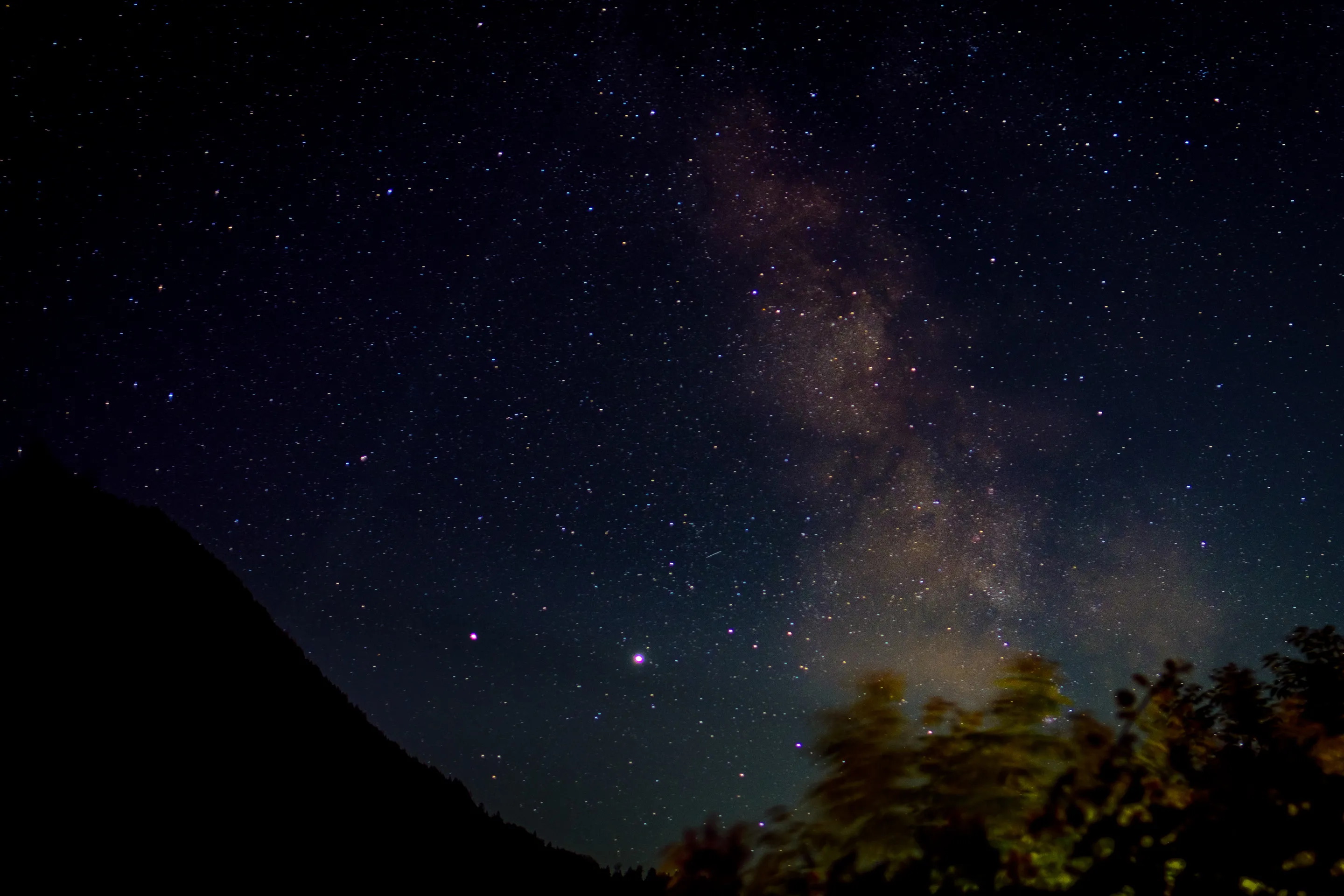 Starry night sky with Milky Way galaxy, mountains and trees silhouetted.