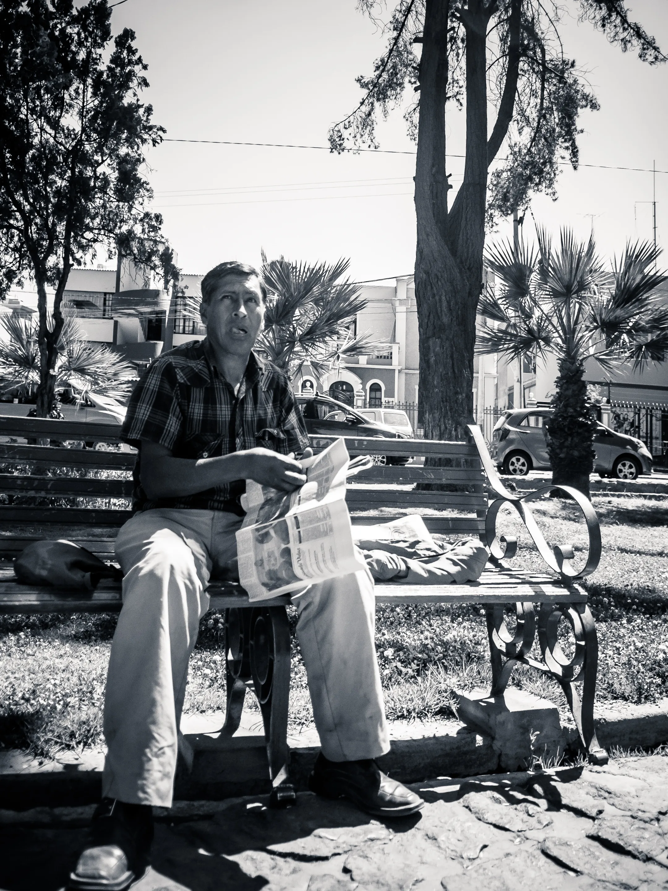 Man sitting on bench in park, reading newspaper, trees and cars in background, black and white photograph.