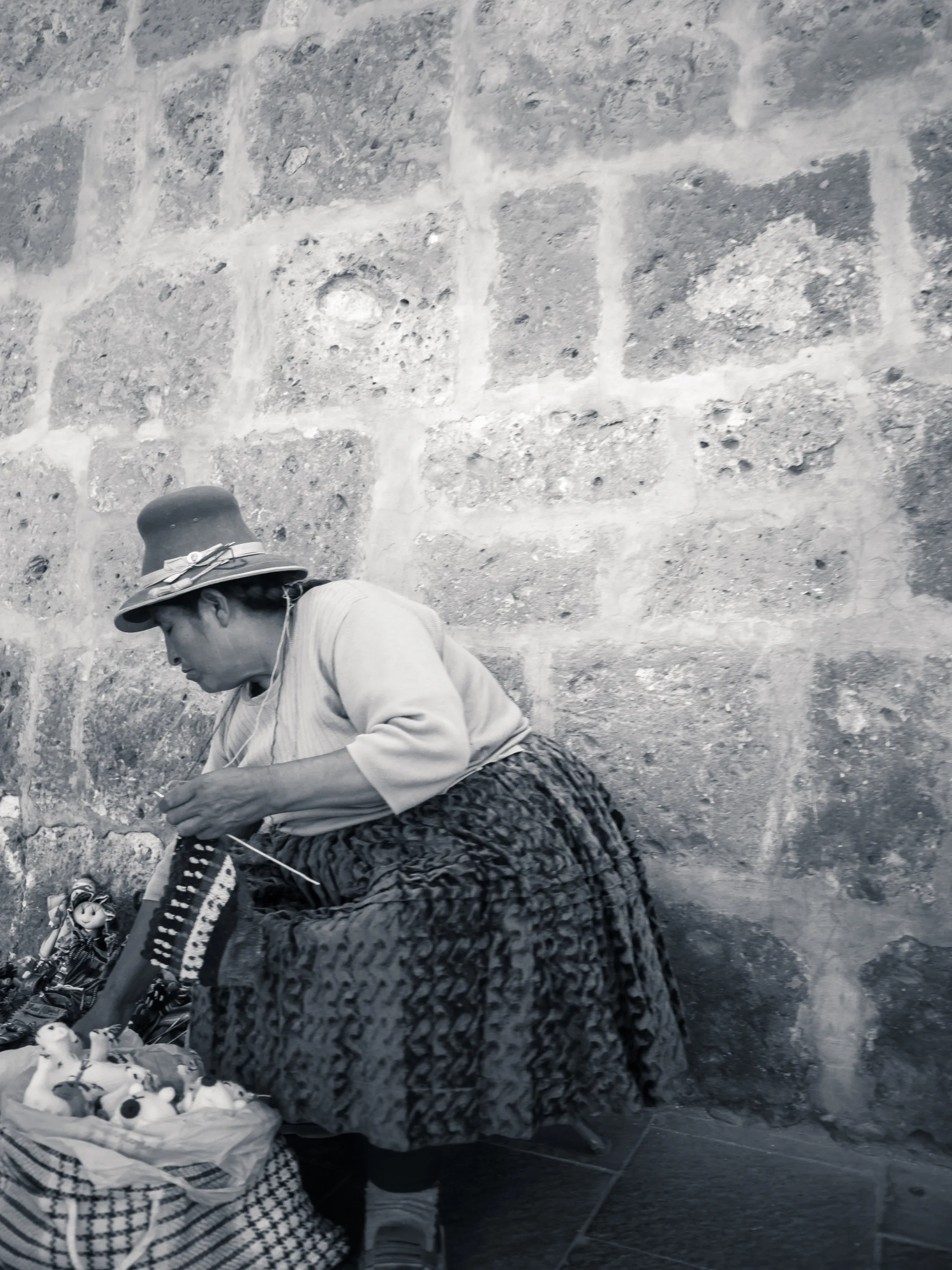 Woman in hat, leaning against stone wall, selling flowers from basket at her feet.