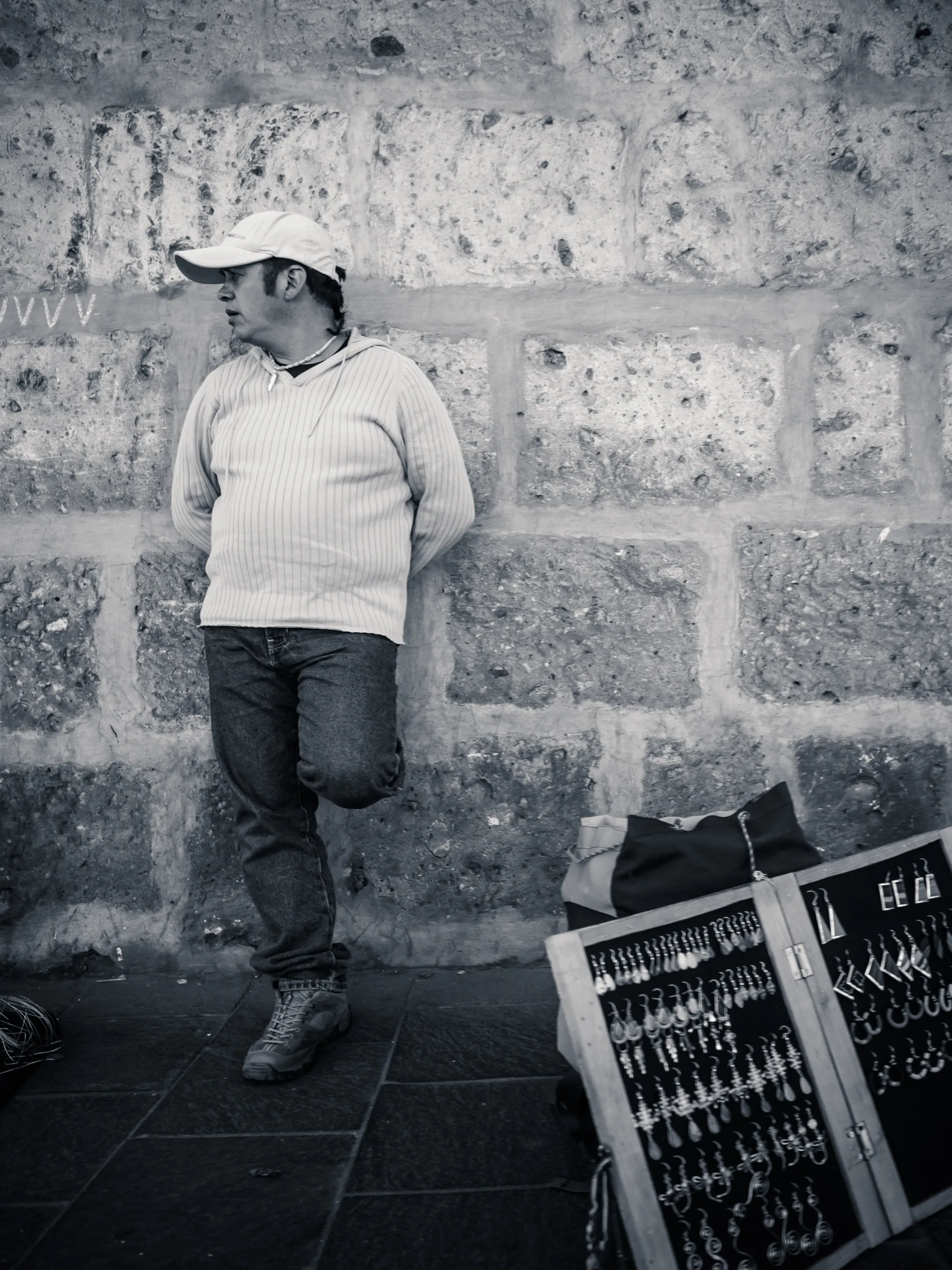 Man in white cap and sweater leaning against brick wall, display of keychains nearby.