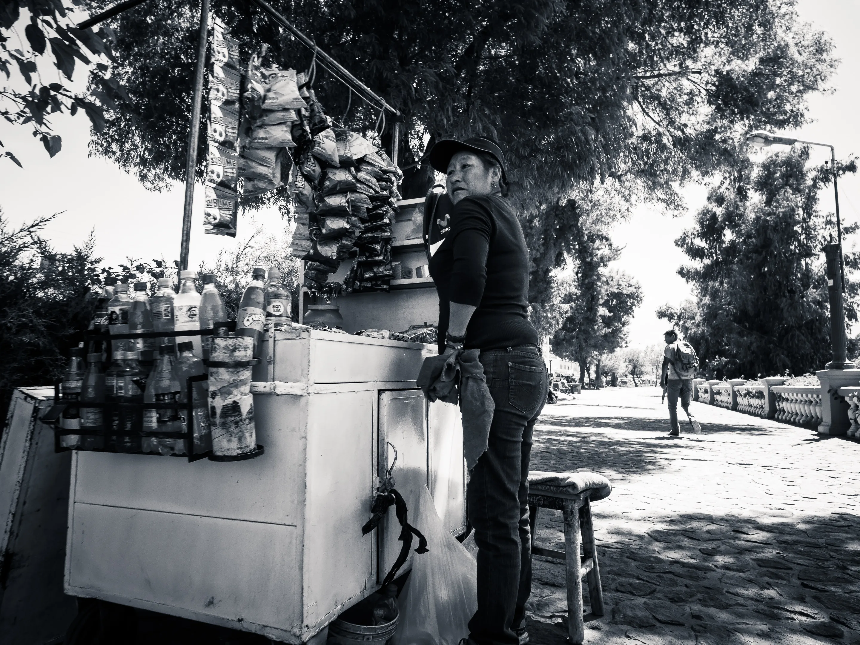 Woman standing beside street food cart with bottles and containers, black and white photo.