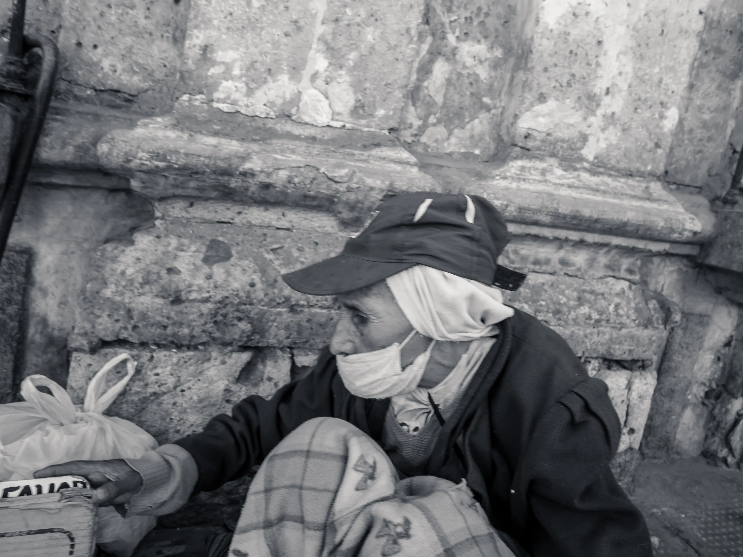 Elderly person wearing a hat and scarf, sitting against a stone wall with bags nearby.