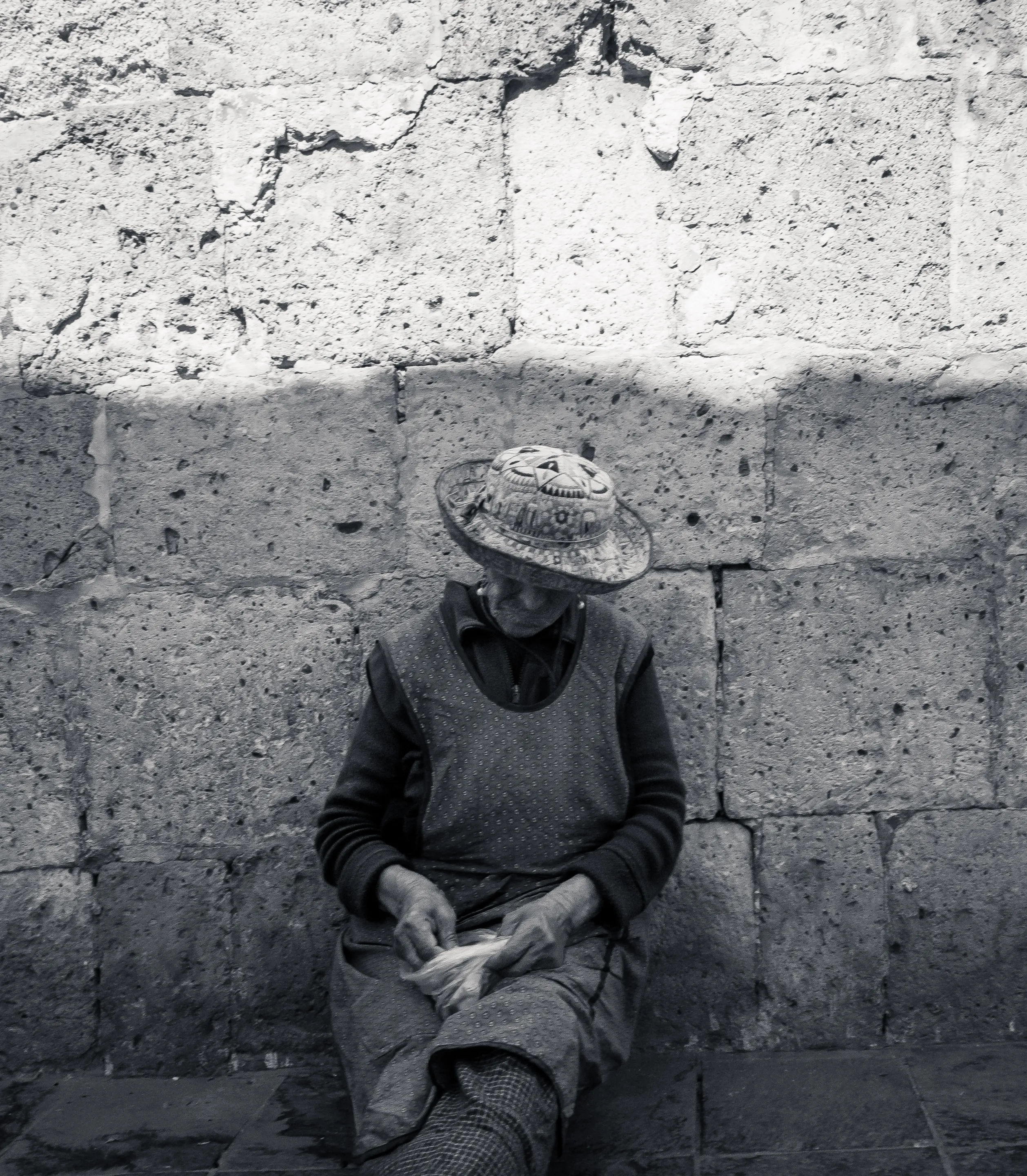 Old woman with hat sitting against stone wall, holding a small object.