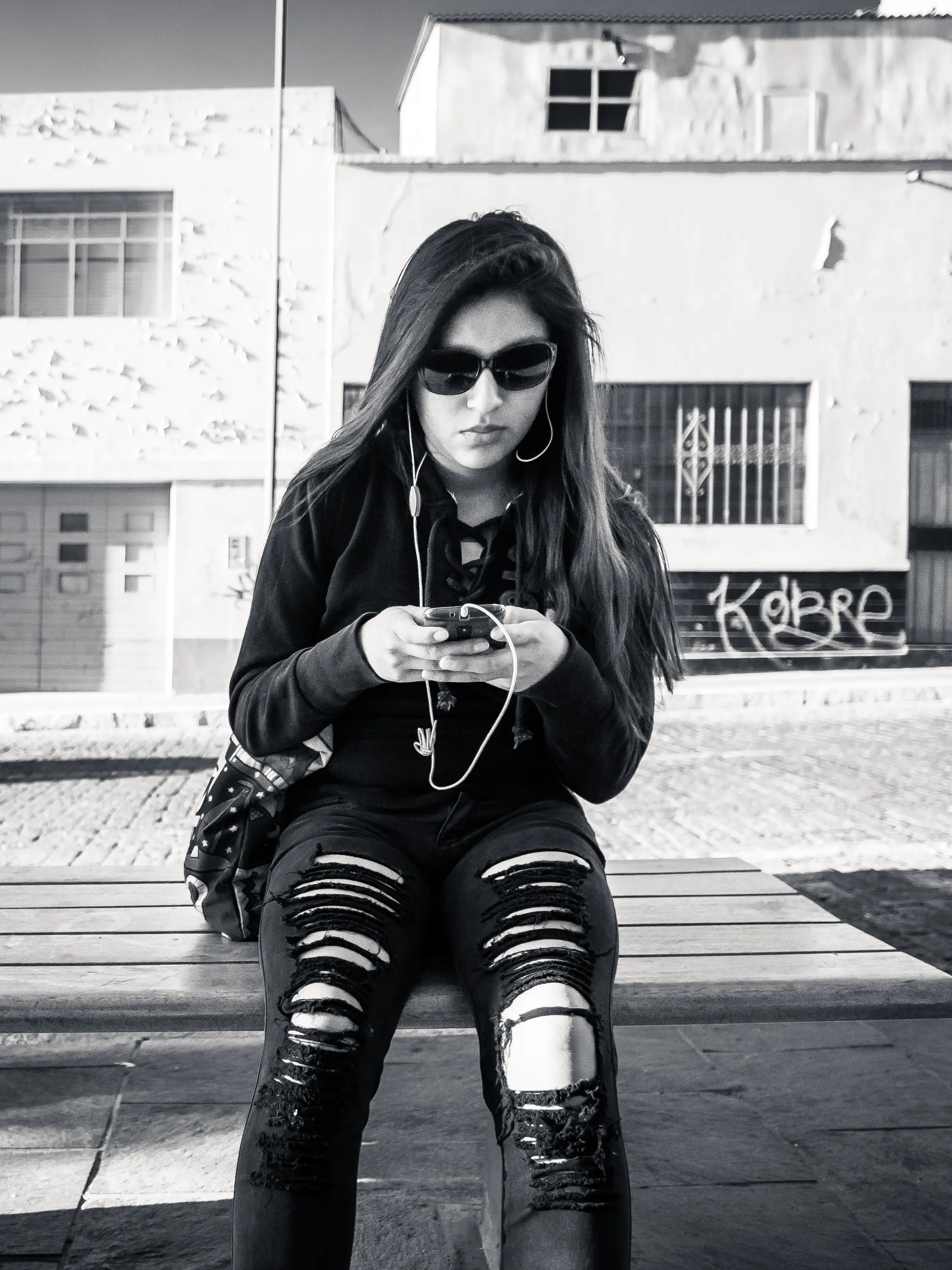 Woman in sunglasses on a bench, using her phone, wearing ripped jeans and boots, against a graffitied wall.